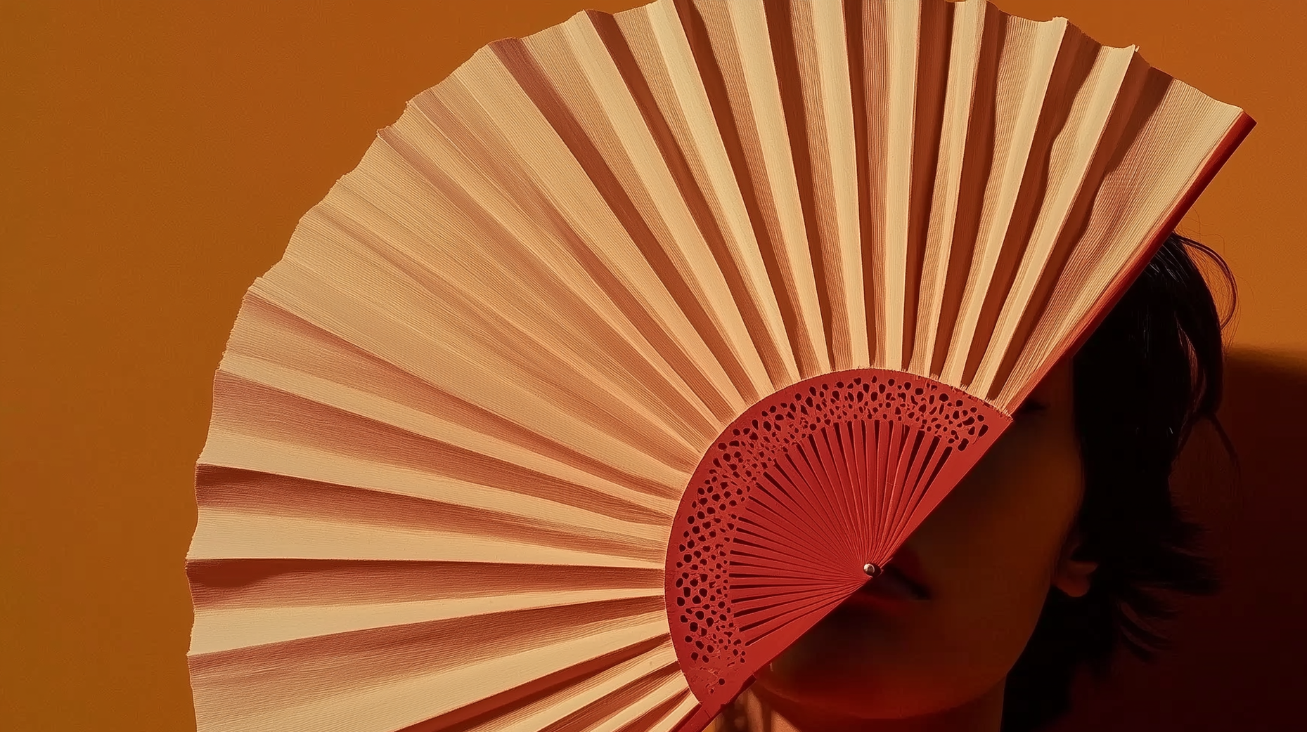 Person holding a large pink folding fan partially covering their face, with dark hair visible against an orange background.