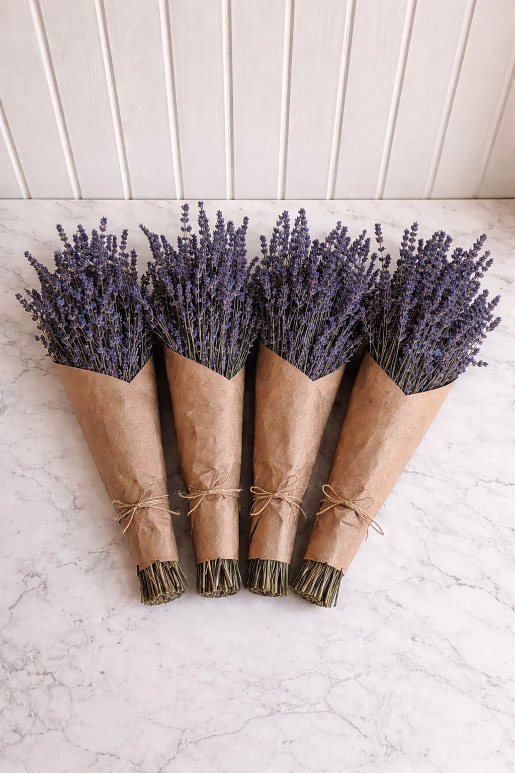 Four bouquets of lavender flowers wrapped in brown paper and tied with string, placed on a white marble surface.