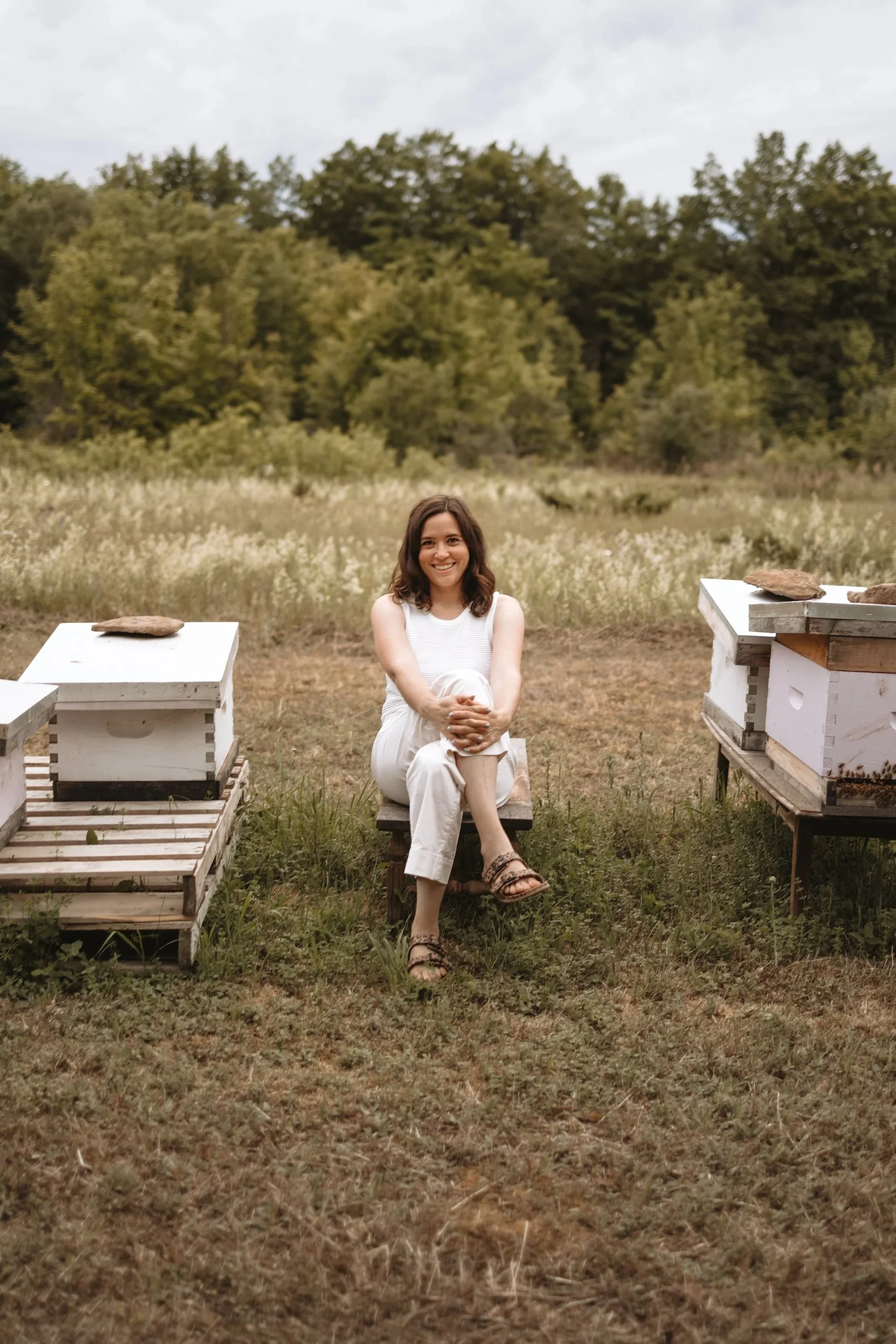 A woman sitting cross-legged on a small wooden chair outdoors, surrounded by beekeeping equipment, with trees and grass in the background.