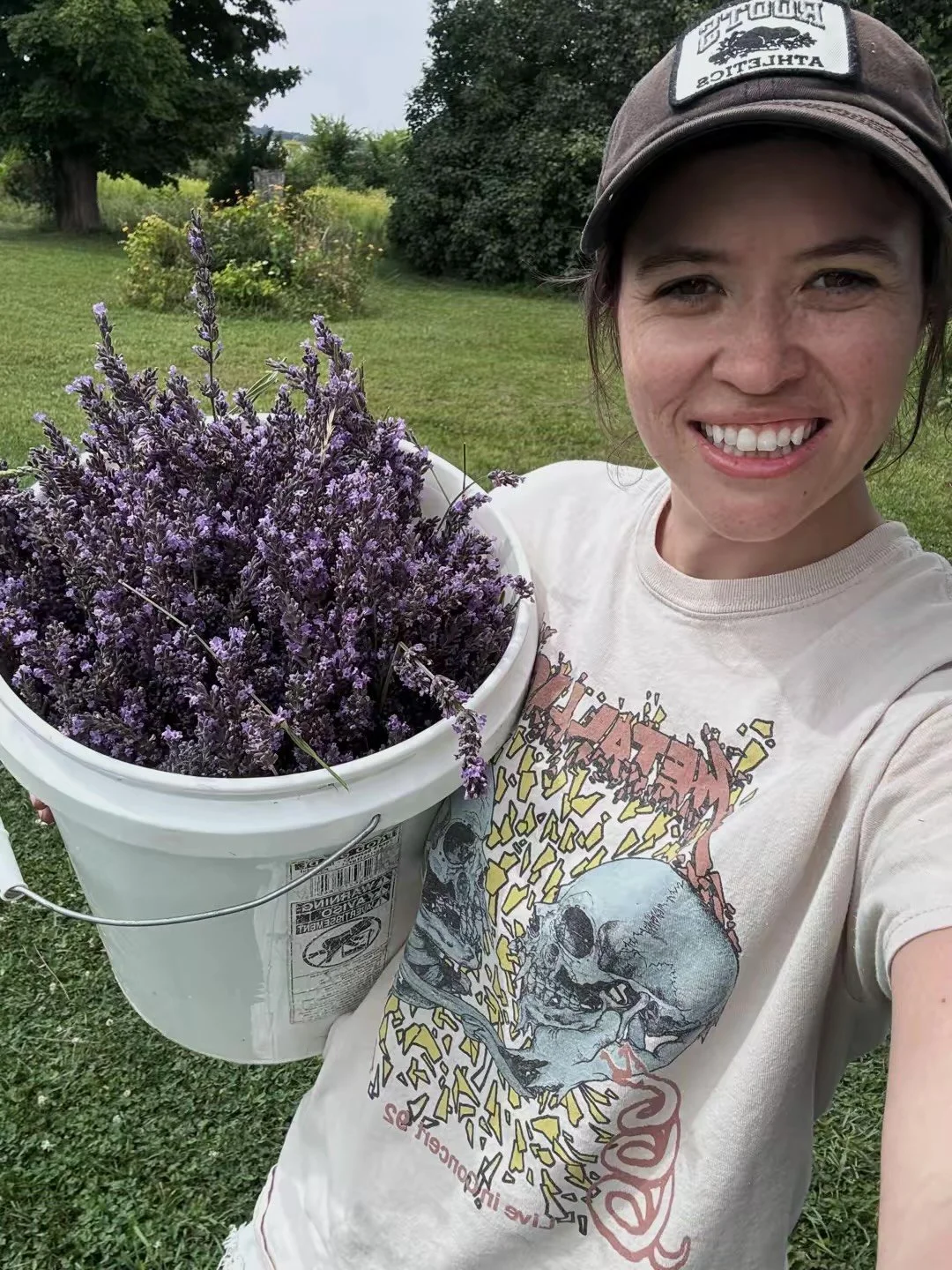 Young woman smiling outdoors while holding a white bucket filled with freshly picked purple lavender flowers.