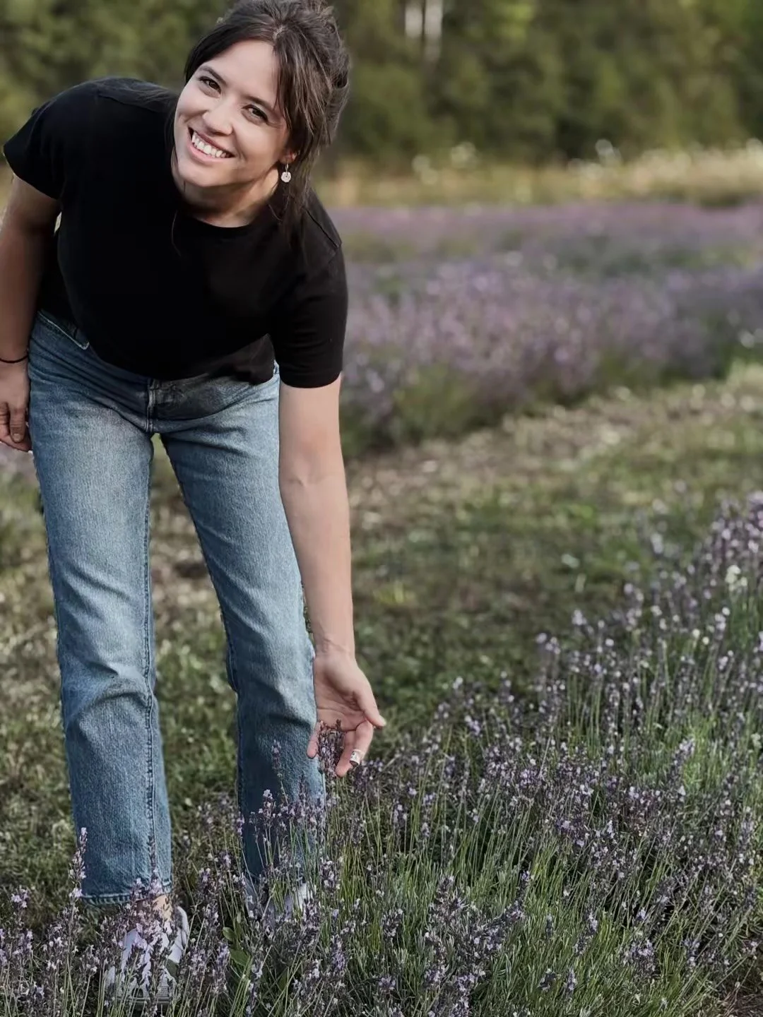 A woman showeing her lavender field, smiling and reaching down to touch the lavender flowers.