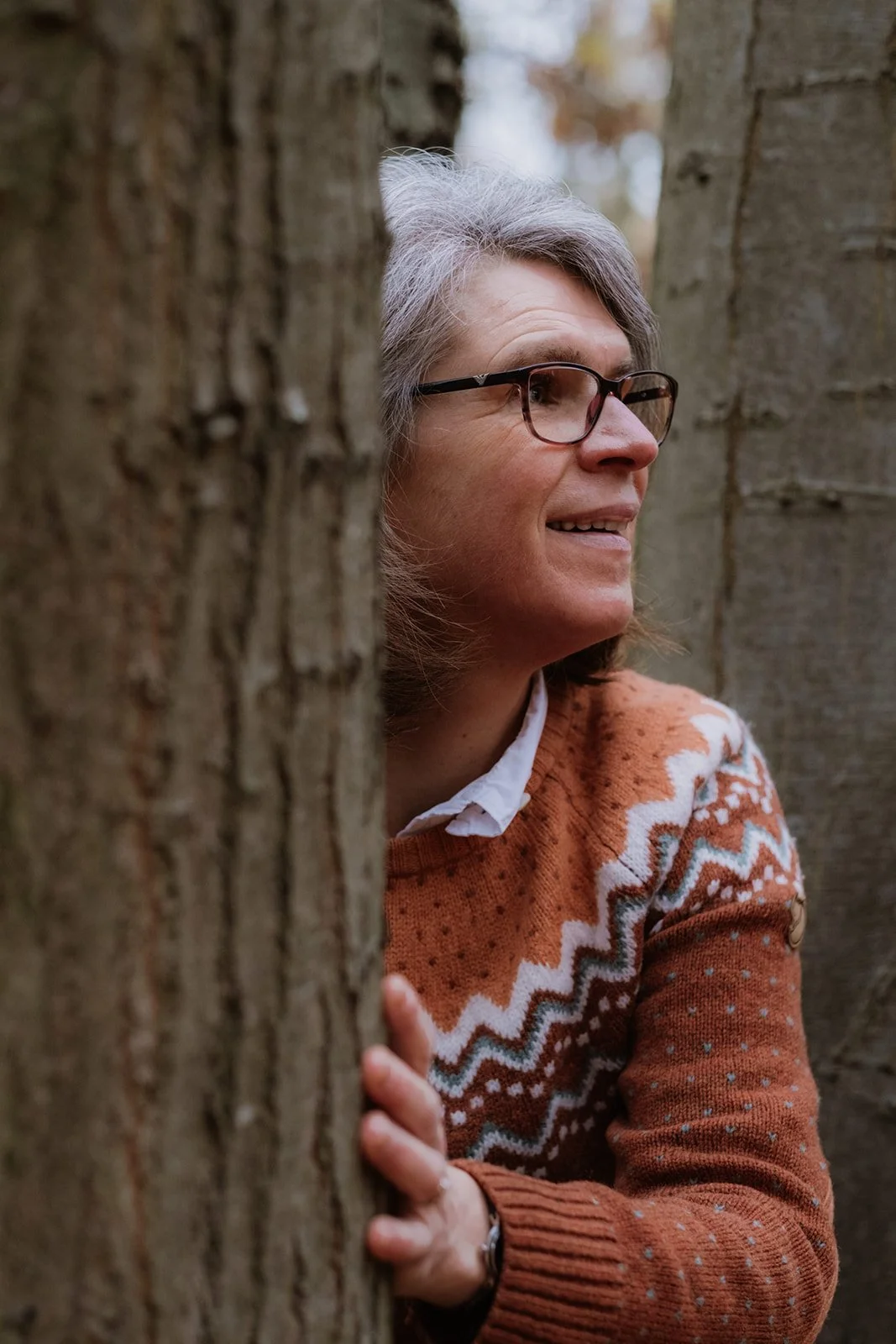 Een vrouw met grijs haar en bril kijkt nieuwsgierig tussen twee bomen door.