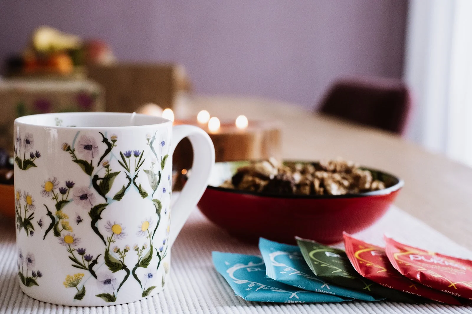 Een witte mug met bloemenpatroon, een rode kom met noten, en verpakkingen met theezakjes op een tafel.