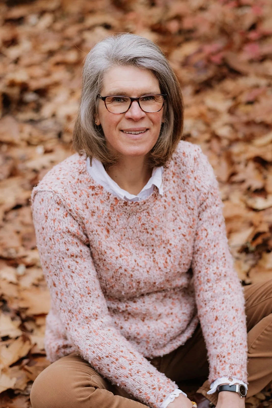 Een vrouw met grijs haar en bril zit op de grond tussen herfstbladeren, draagt een roze-oranje gebreide trui en bruin broek, lachend naar de camera.