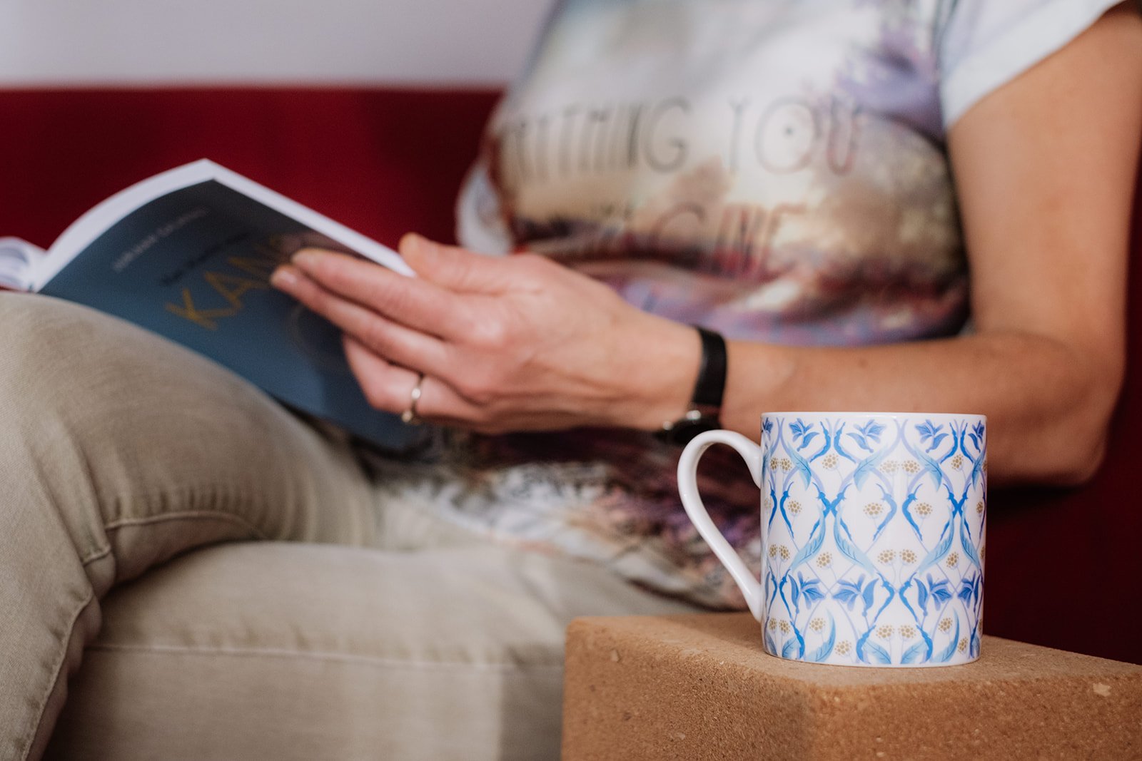 Persoon zit op de bank, leest een boek met blauwe en gouden letters, een koffie- of theemok met blauw en gouden patroon staat op een blokje.