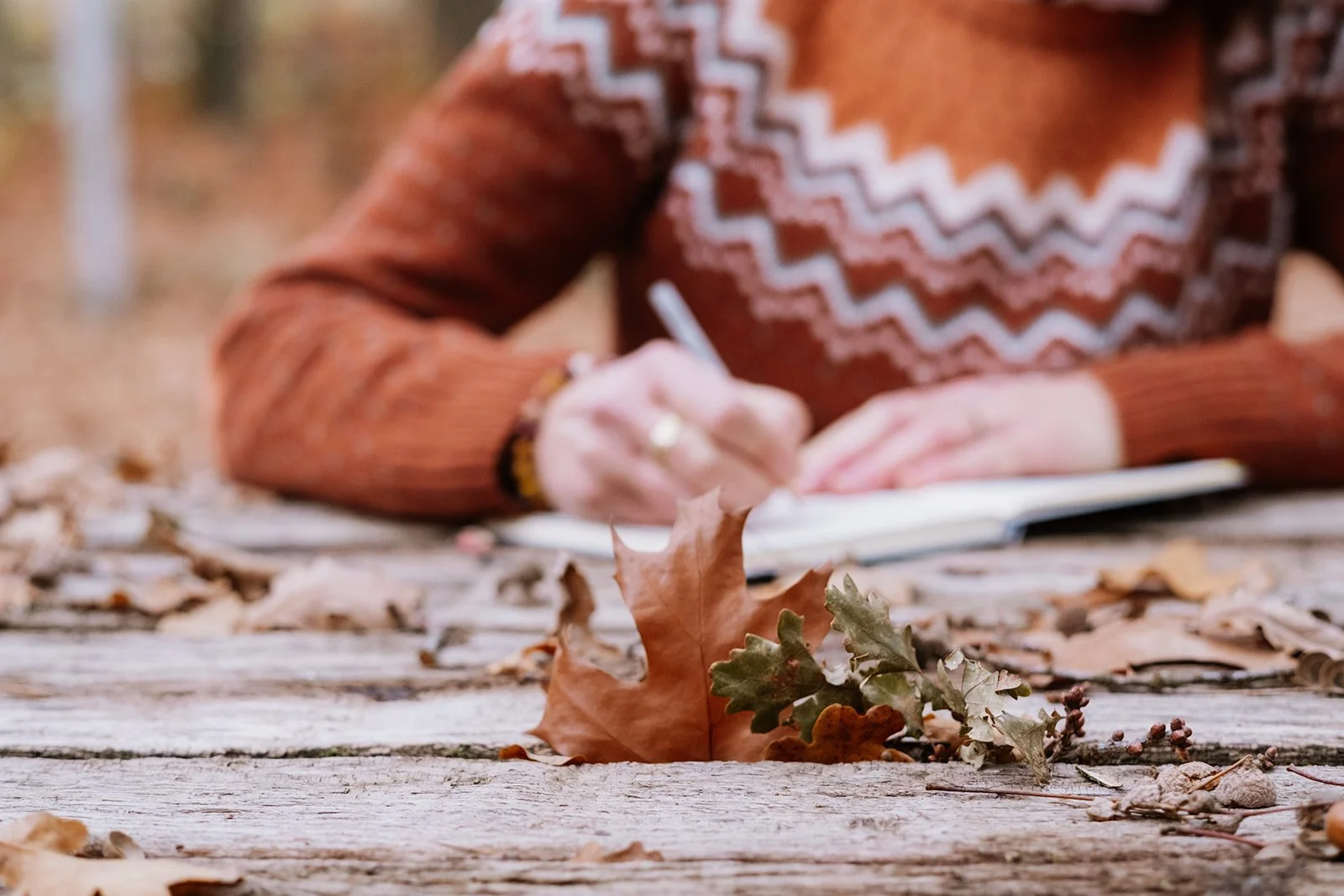 Vrouw schrijft in een notitieboekje op een houten tafel met herfstbladeren en eikenbladeren in de voorgrond, herfstbos op de achtergrond.