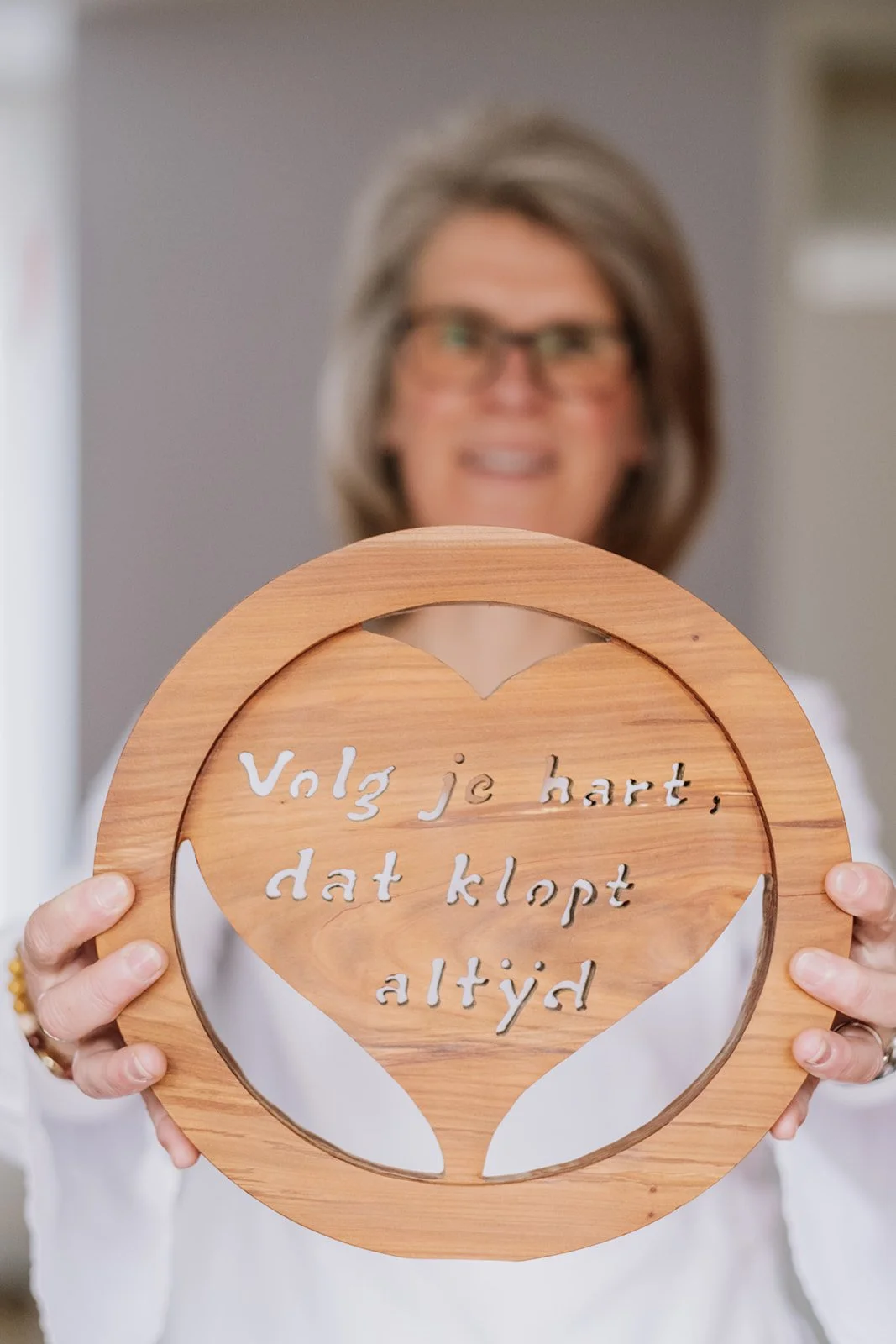 Een vrouw houdt een houten bord met de tekst 'Volg je hart, dat klopt altijd' in een hartvorm in de voorgrond, terwijl ze op de achtergrond lachend zichtbaar is.