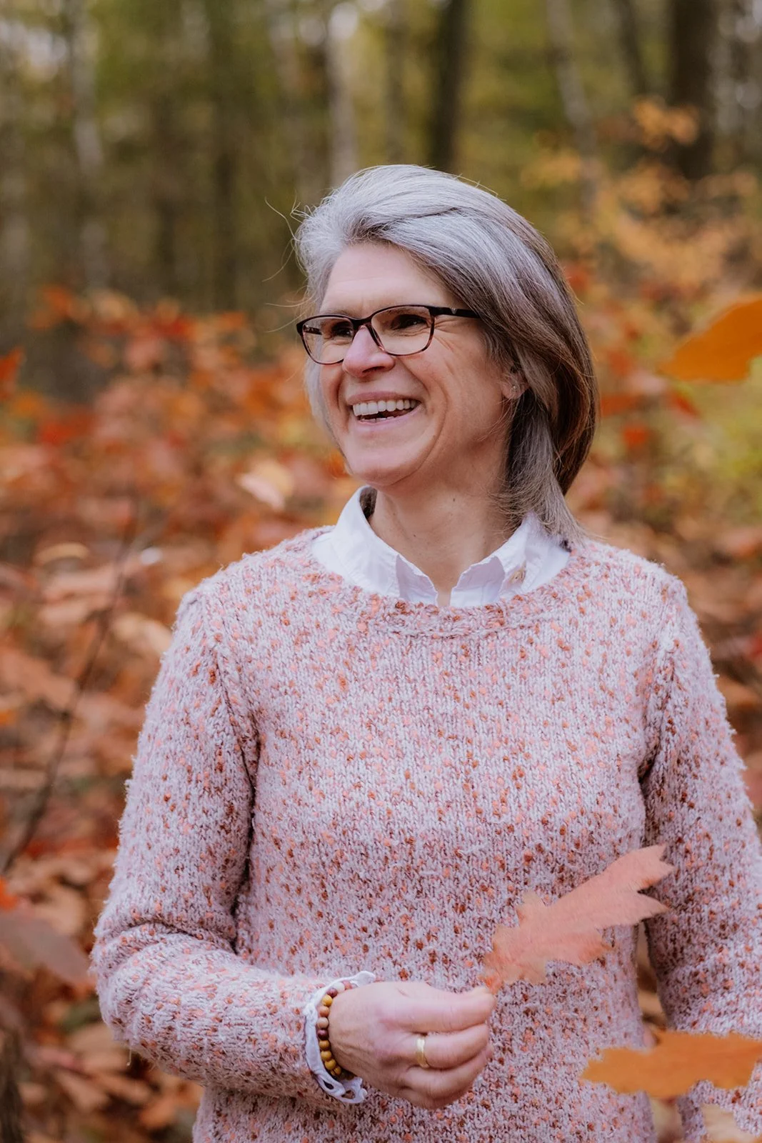 Een lachende vrouw met grijs haar en een bril, die in een herfstbos staat, terwijl ze een herfstblad vasthoudt.