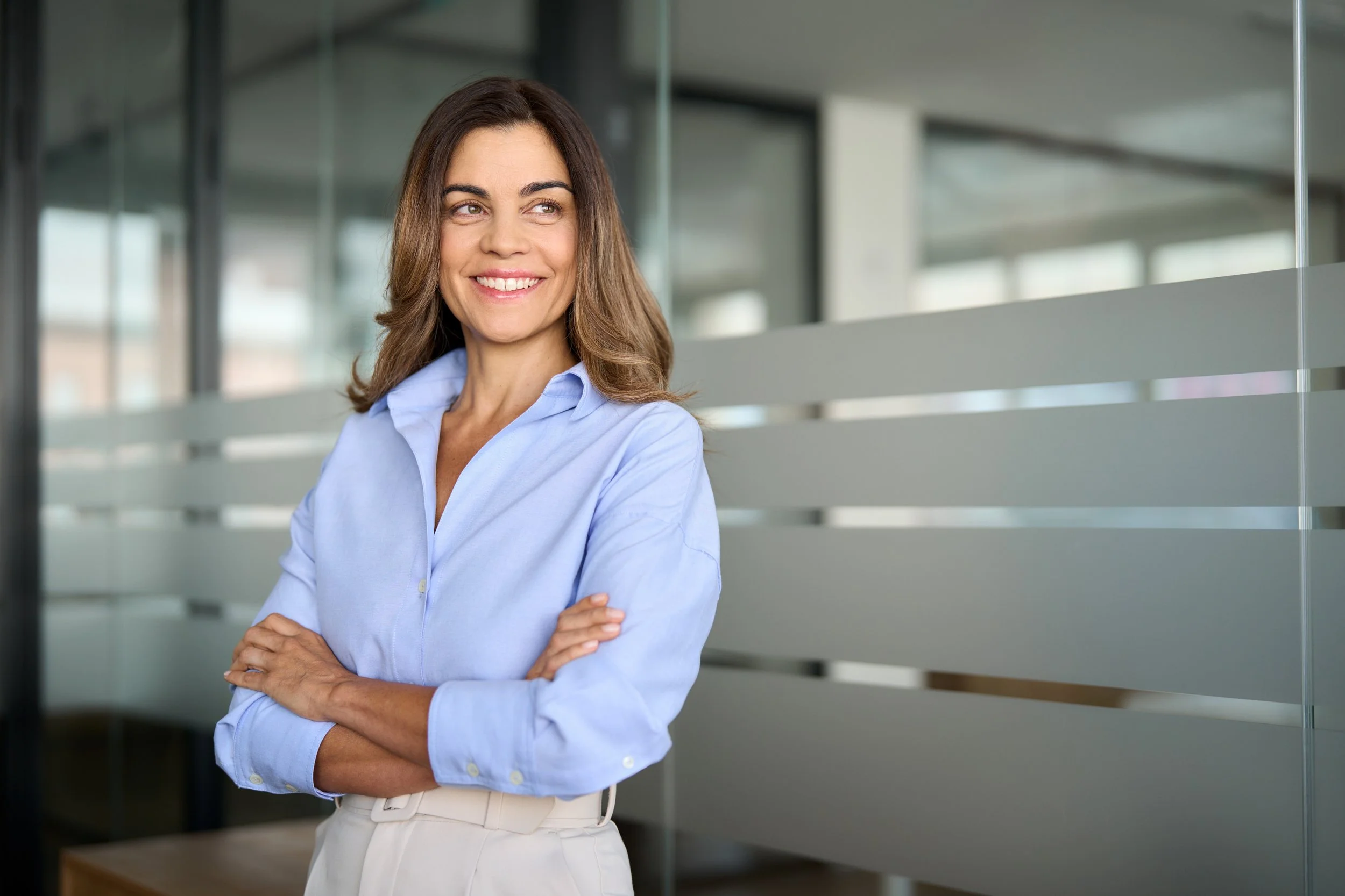 A woman in a light blue button-up shirt standing in front of a modern office interior with glass walls, smiling confidently with arms crossed.