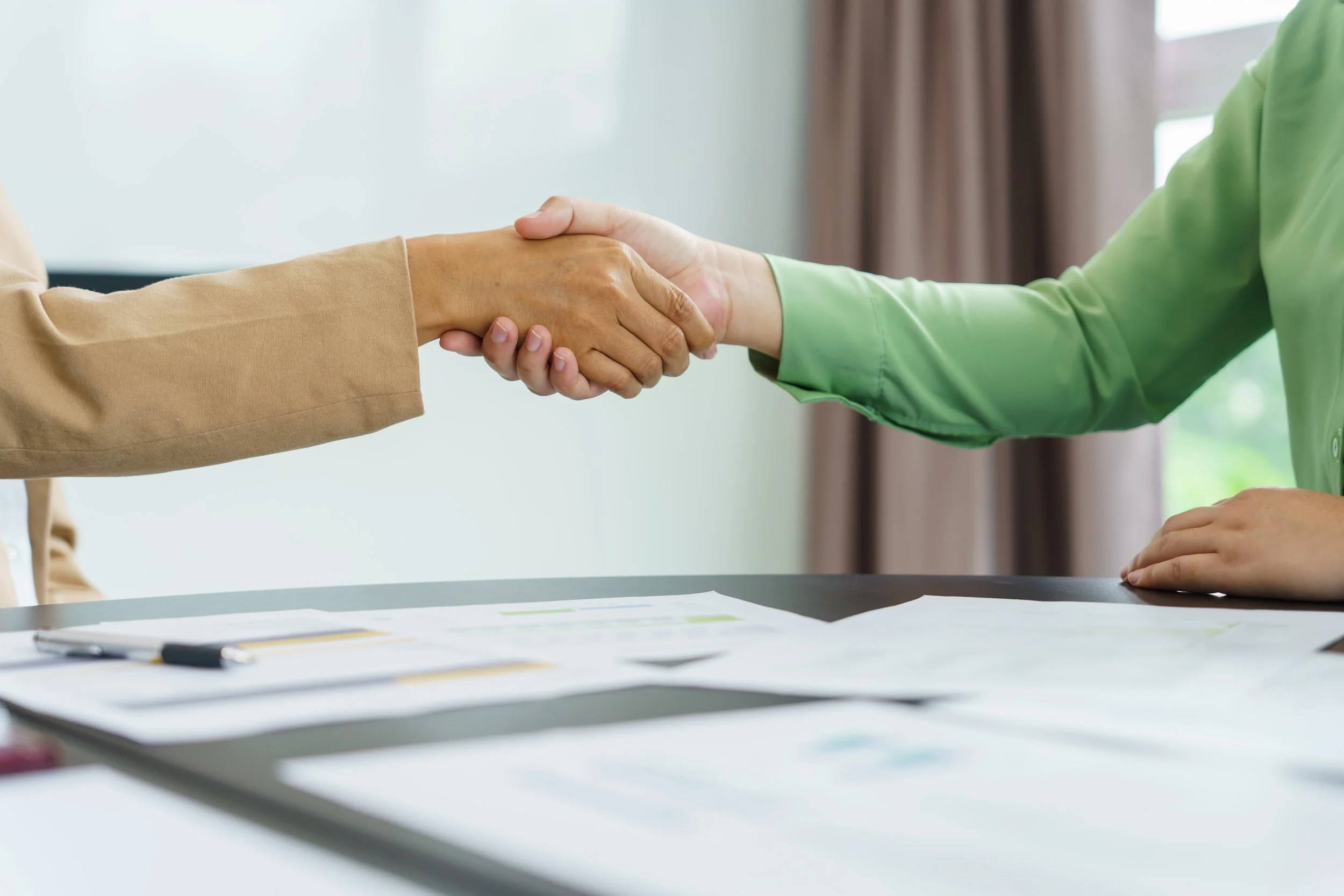 Two people shaking hands over a desk with papers and a pen, in a professional setting.