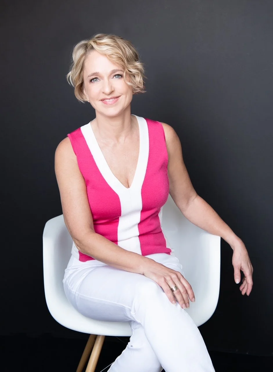 A woman with short blonde hair sitting on a white chair in front of a gray background, wearing a pink sleeveless top with white accents and white pants, smiling softly at the camera.