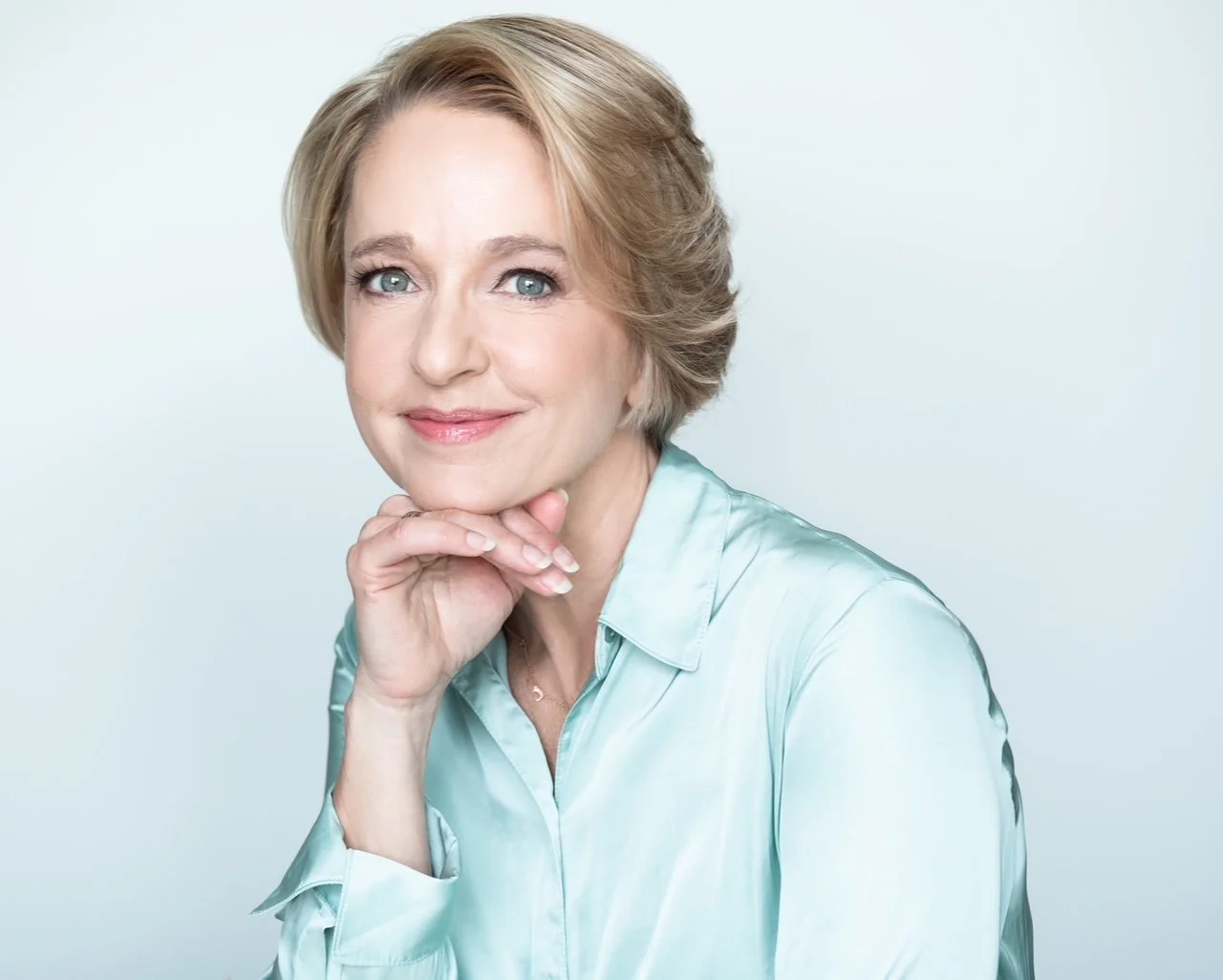 A middle-aged woman with short blonde hair and blue eyes, smiling gently with her chin resting on her hand, wearing a light blue silky blouse against a plain light background.