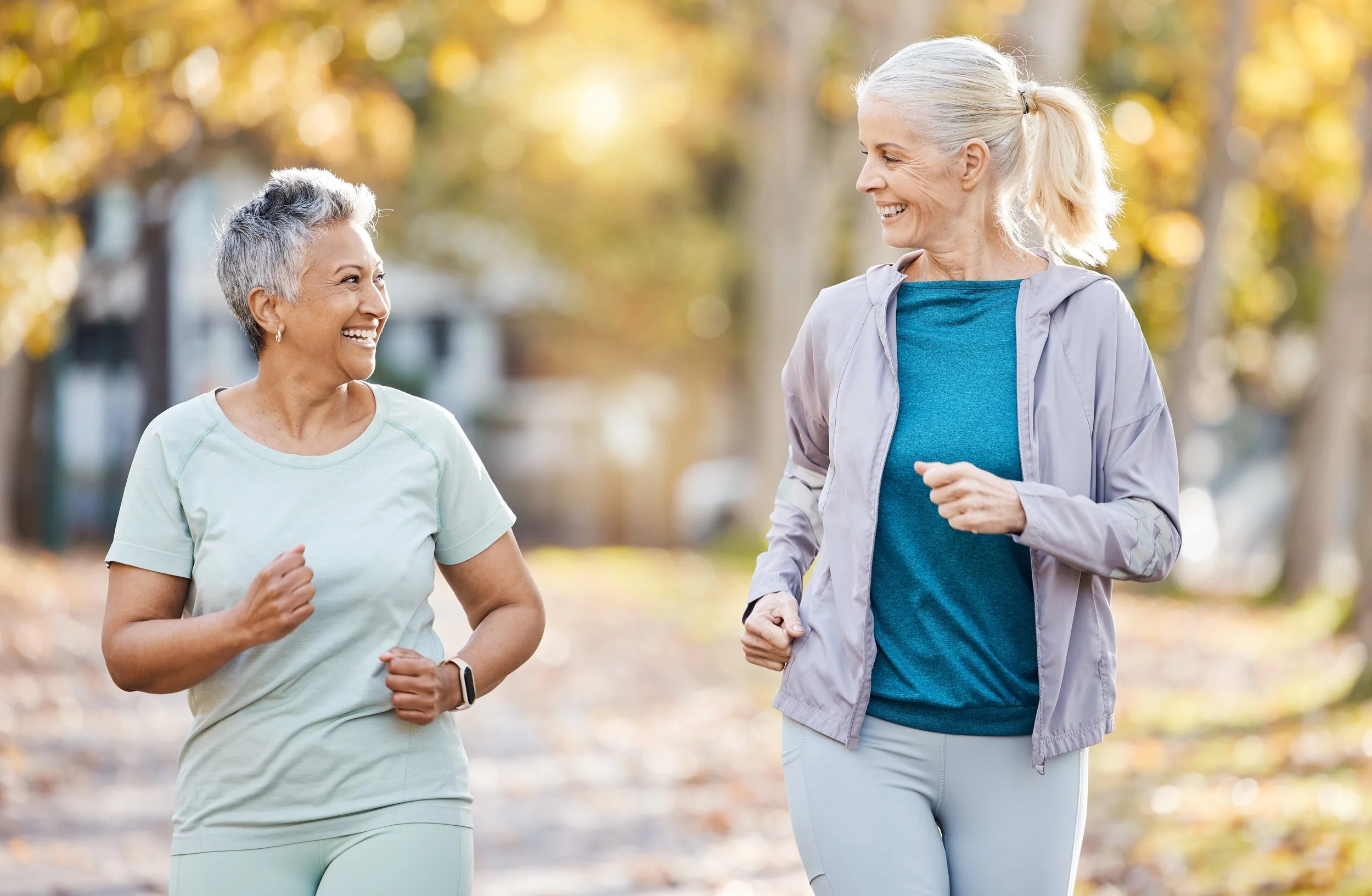 Two older women jogging together outdoors on a fall day, smiling and looking at each other.