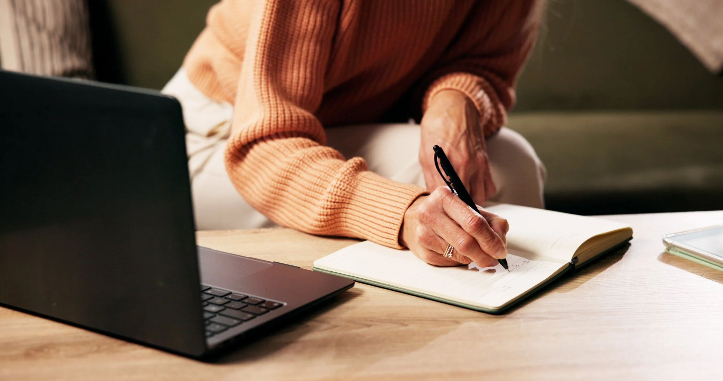 Person writing in an open notebook with a pen, sitting at a desk with a laptop and a tablet nearby.