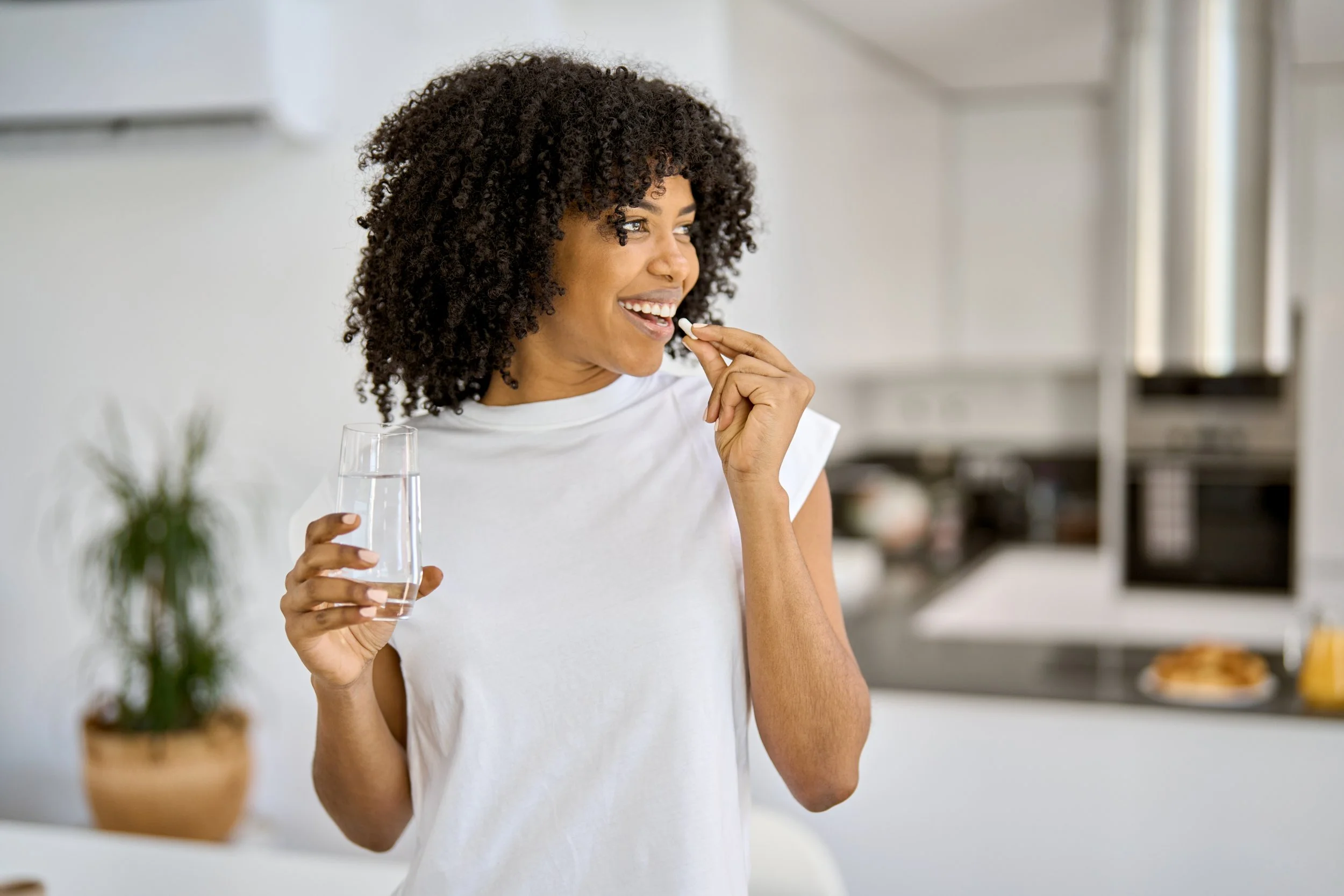 A woman with curly hair holds a glass of water in one hand and takes a pill with the other hand, standing in a bright kitchen.