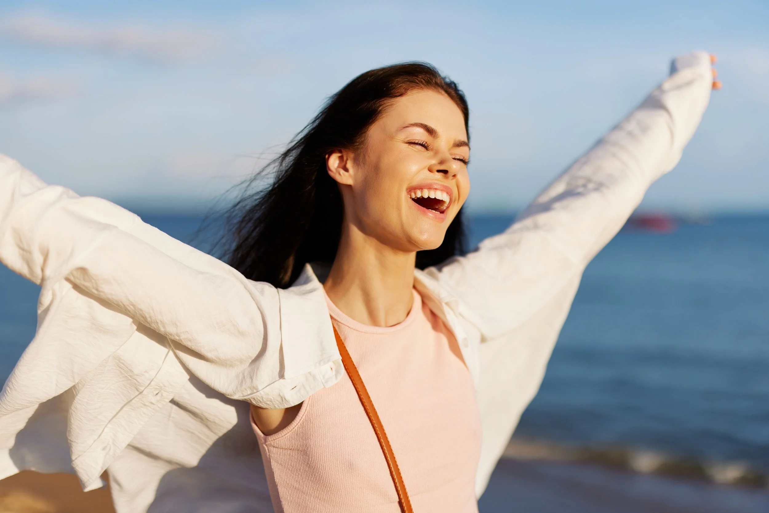 A woman joyfully celebrates at the beach with arms outstretched, smiling with closed eyes, near the ocean.
