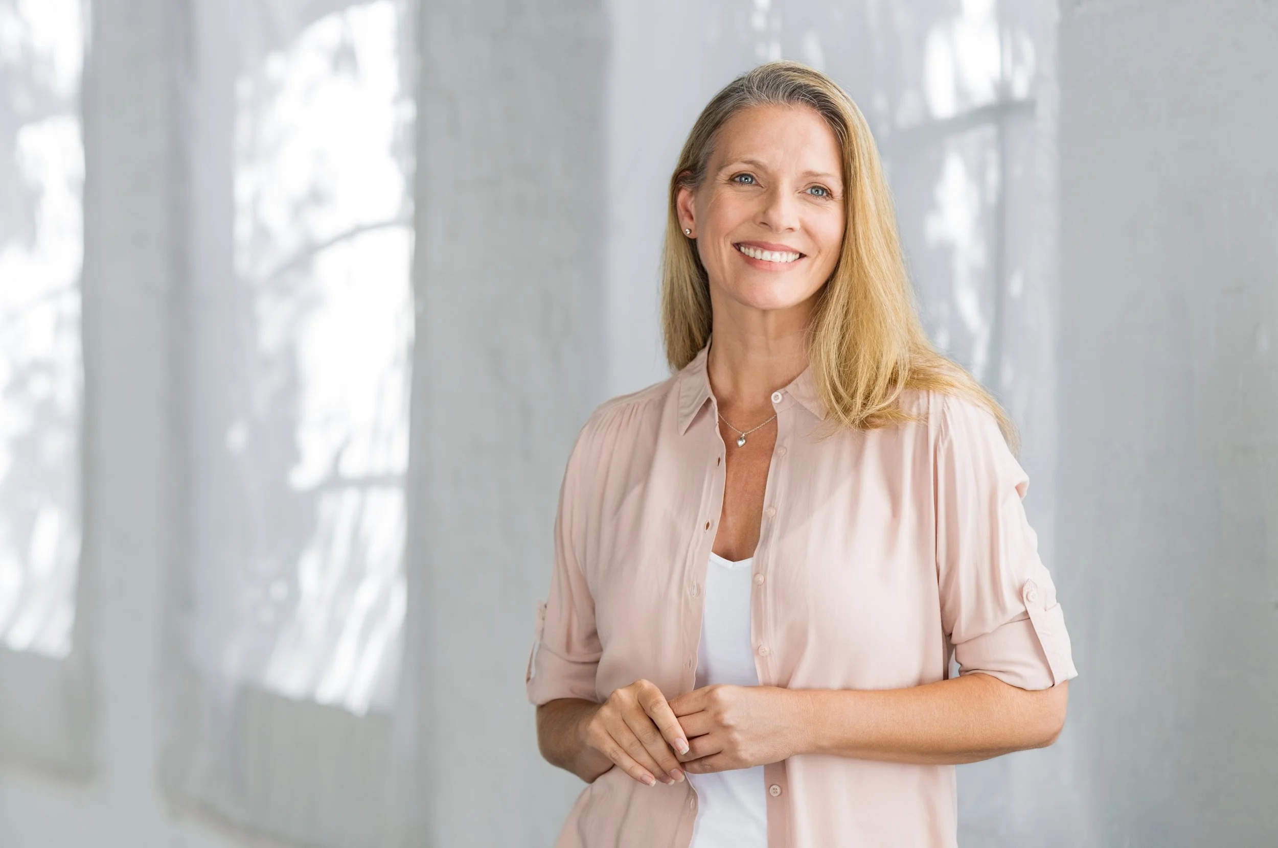 A smiling middle-aged woman with blonde hair wearing a light pink shirt and jewelry, standing indoors near windows.