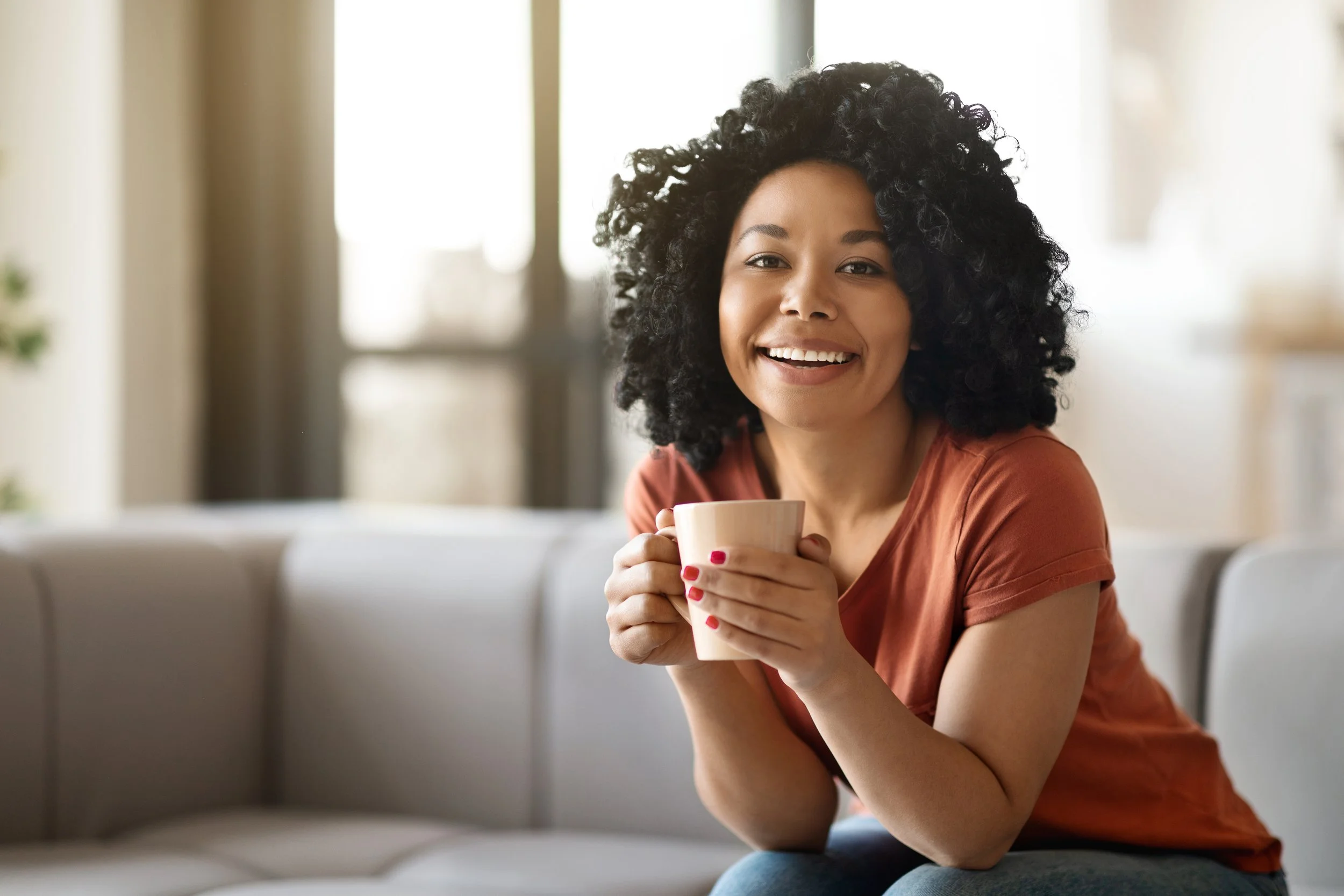 A woman with curly black hair, smiling and holding a beige mug, sitting on a light-colored sofa in a bright room with large windows.