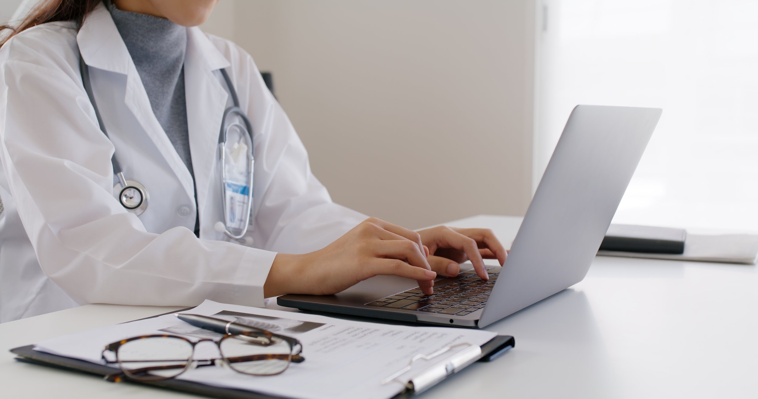 A healthcare professional in a white lab coat with a stethoscope around their neck, using a laptop at a desk with papers and glasses.