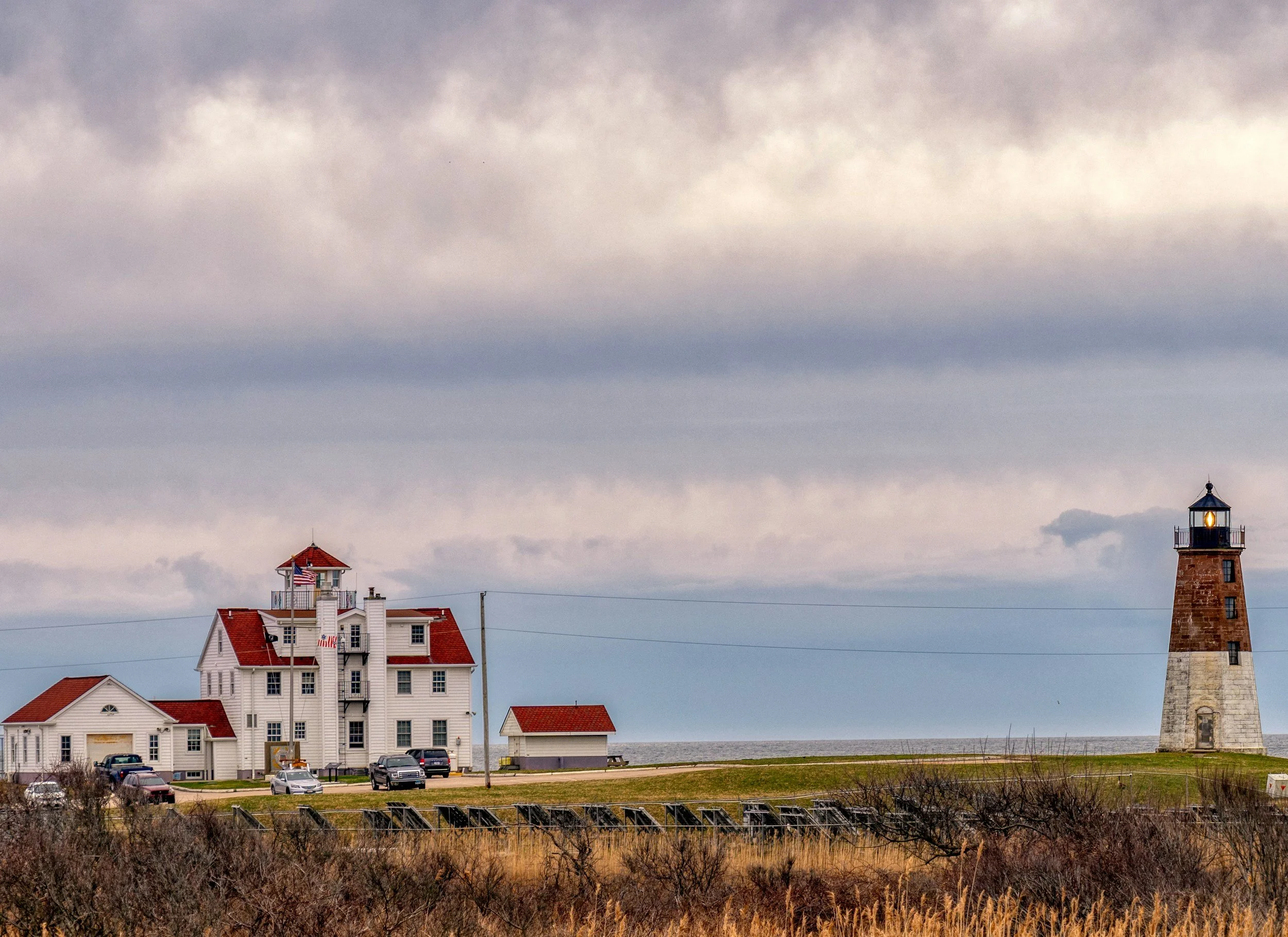 A lighthouse on the right side of the image, a white building with a red roof and a smaller white building on the left, both near the beach with a few parked cars in front, and a cloudy sky above.