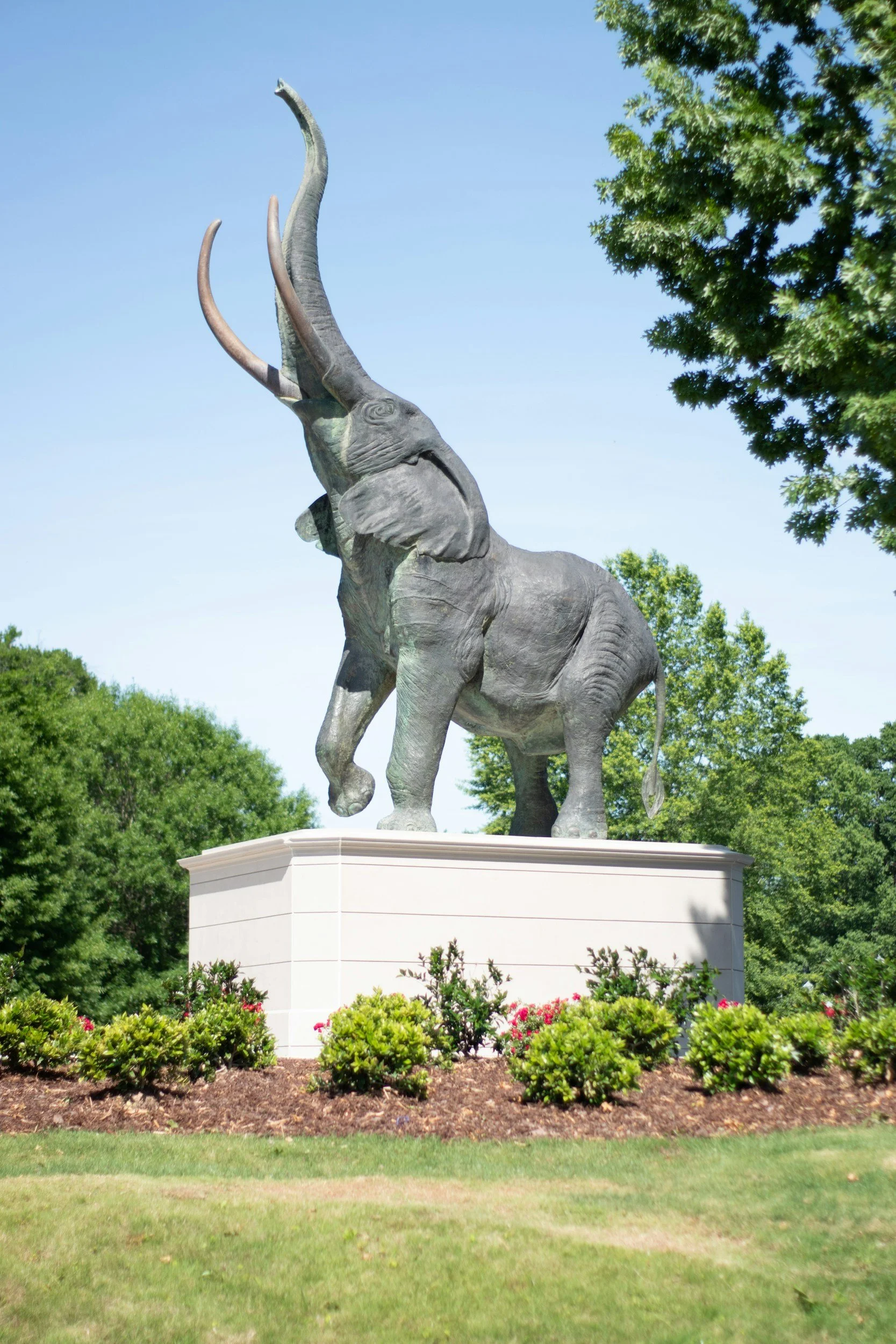 A large outdoor statue of an elephant with its trunk raised, situated on a white pedestal, surrounded by landscaped bushes and trees under a clear blue sky.