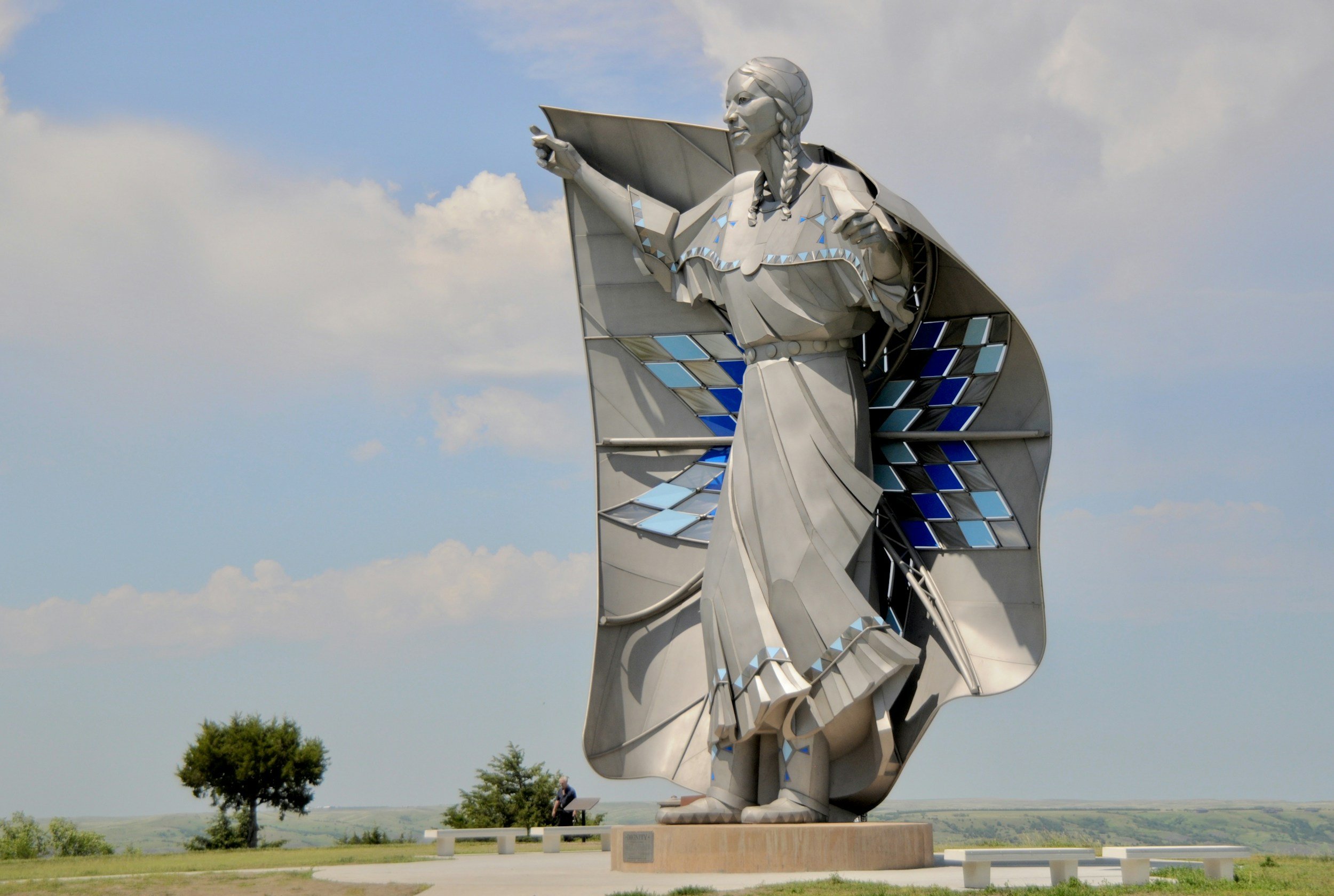 A large metallic statue of a Native American man in traditional dress, with long braided hair, standing outdoors under a cloudy sky, pointing forward with one hand. The statue features geometric blue and silver accents.