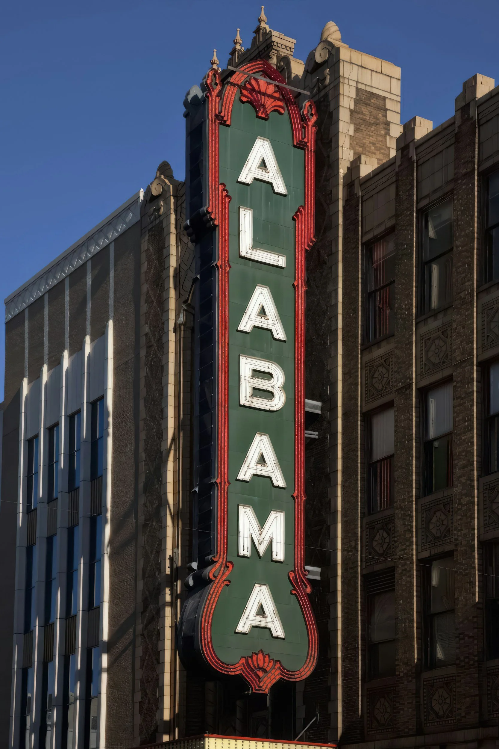 Vertical sign with the word 'ALABAMA' in large white letters on a green background, surrounded by decorative red and black trim, attached to an ornate building facade. Used to illustrate the shop directories for Alabama.