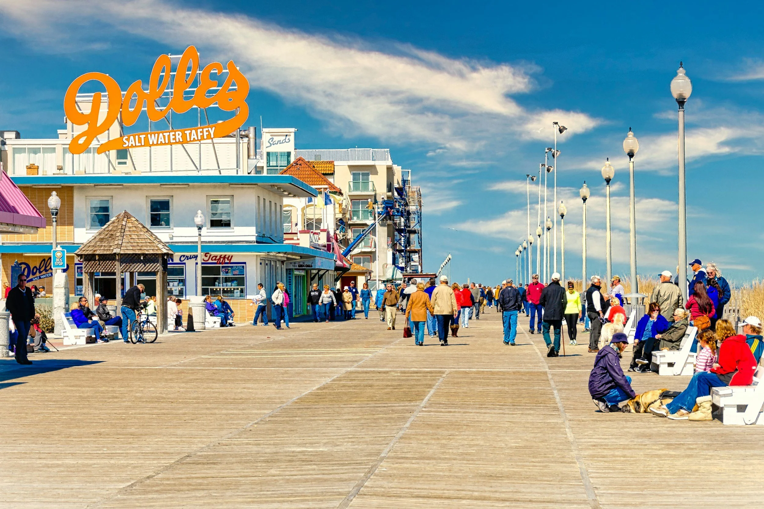 The image shows a busy pier with people walking and sitting on benches on a sunny day. There are colorful shops, street lamps, and a large sign reading 'Galle's Salt Water Taffy'. Used to illustrate the shop directories for Delaware.