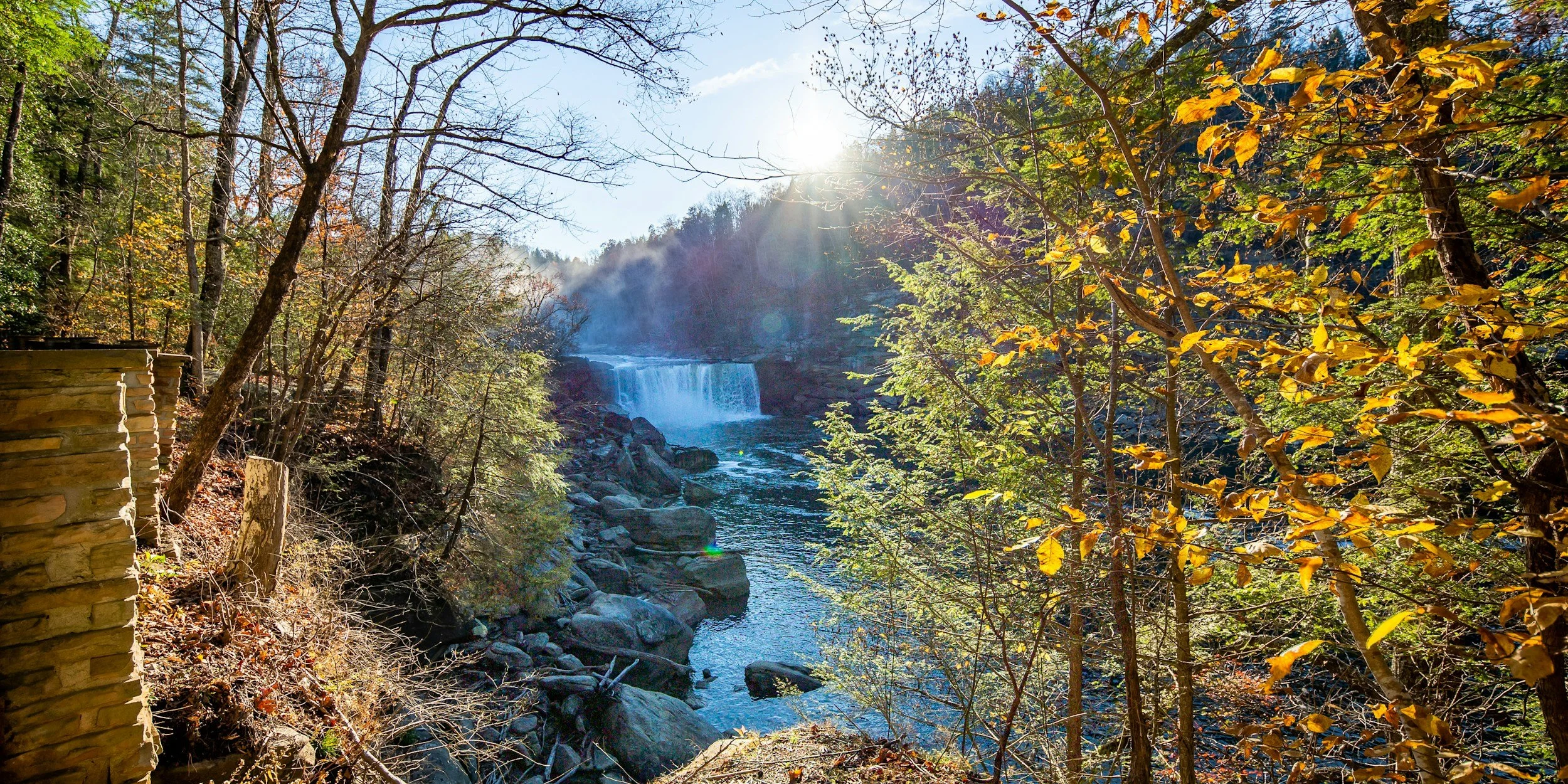 Sunlight filters through trees in a forest near a waterfall cascading over rocks into a stream, with autumn-colored leaves on branches.