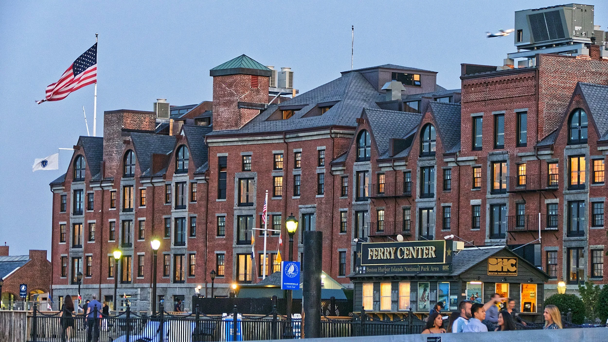 View of a historic brick building with a sign that reads 'FERRY CENTER' in a busy waterfront area at dusk, with people walking and socializing, American flags, street lamps, and a small boat dock visible.