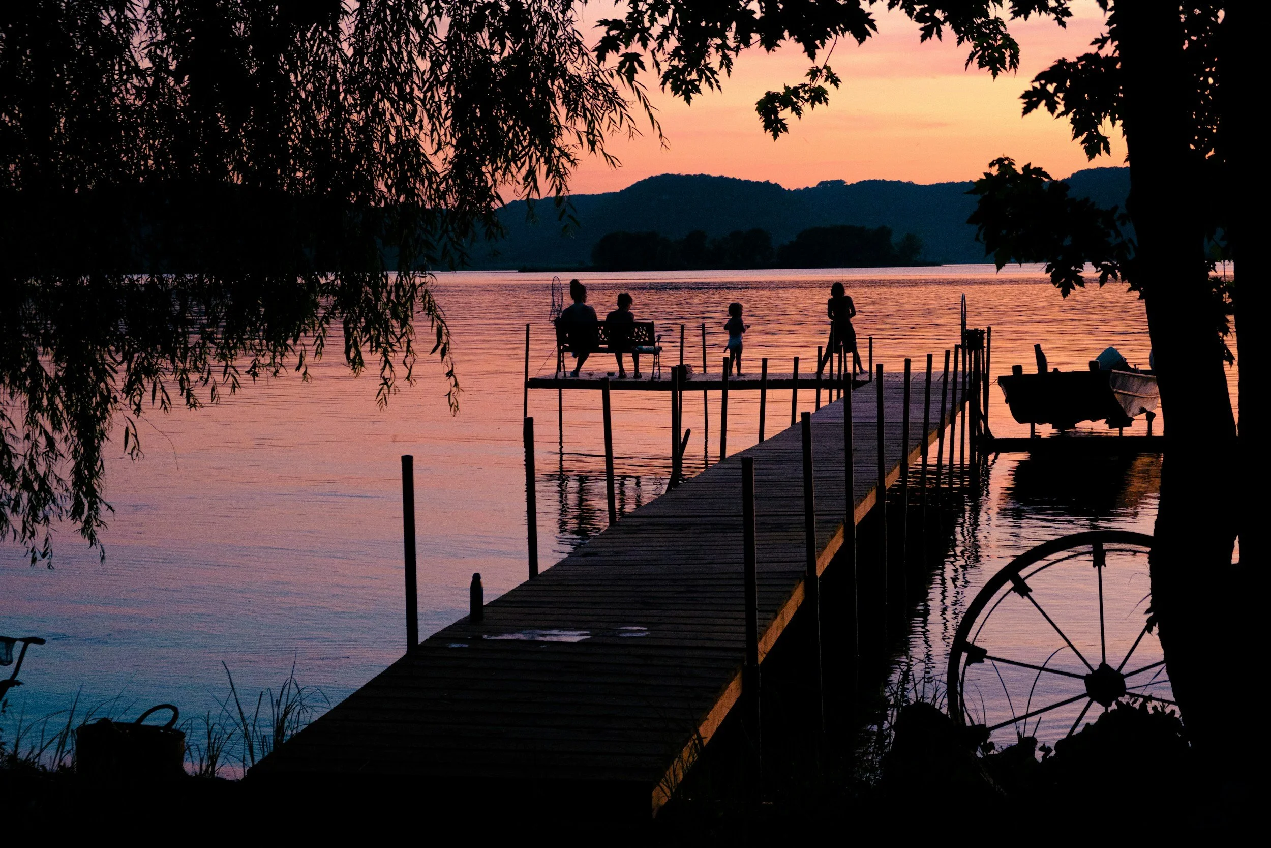 Silhouette of people on a dock during sunset at a lake, with trees framing the scene and mountains in the background.
