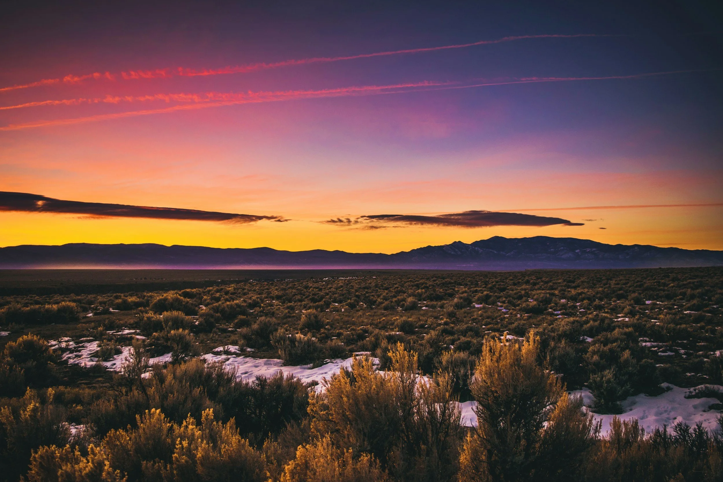 Sunset over a desert landscape with mountains in the distance, sparse bushes, and patches of snow on the ground.