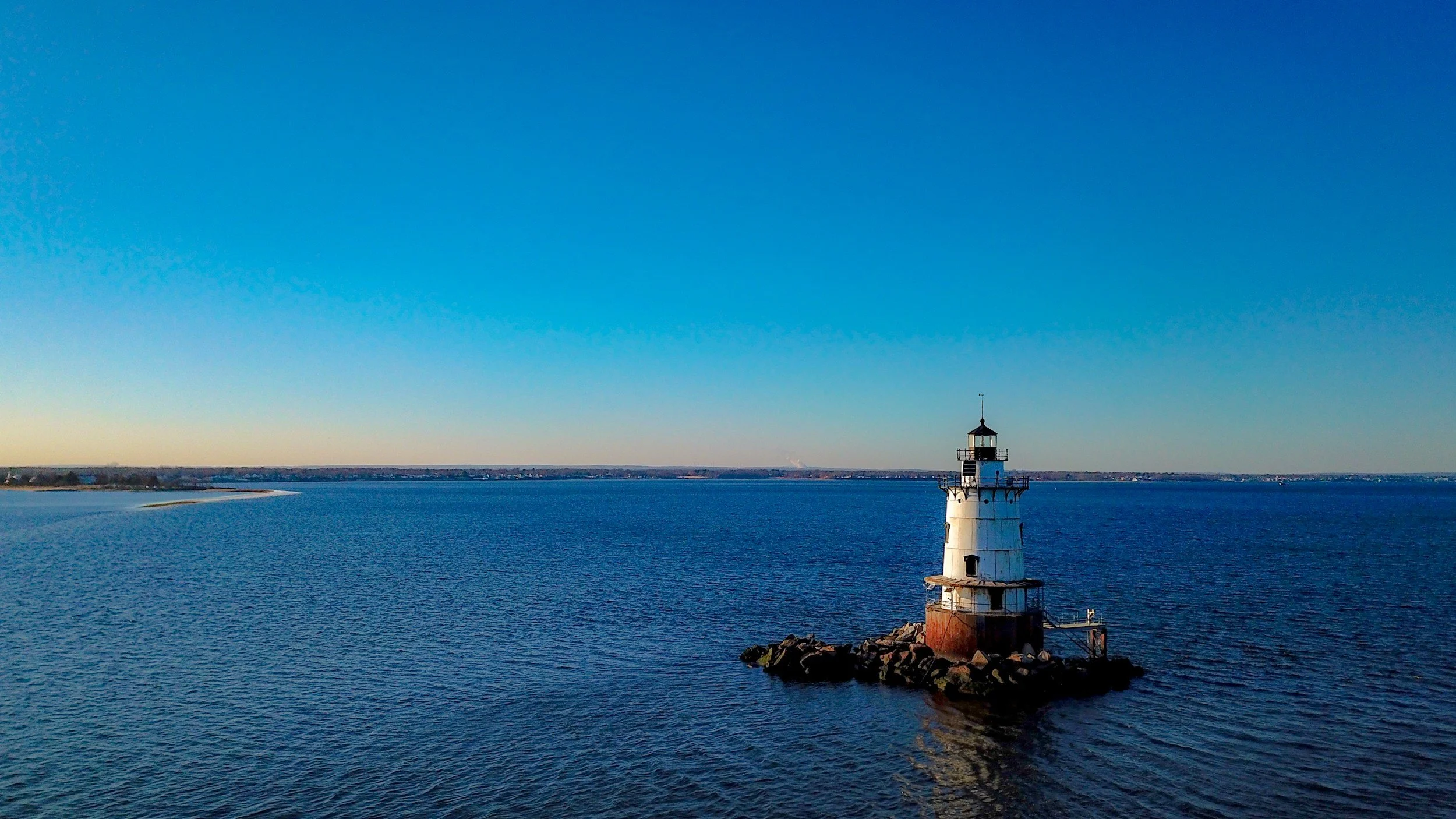 A lighthouse on a small rocky island surrounded by calm water under a clear blue sky.