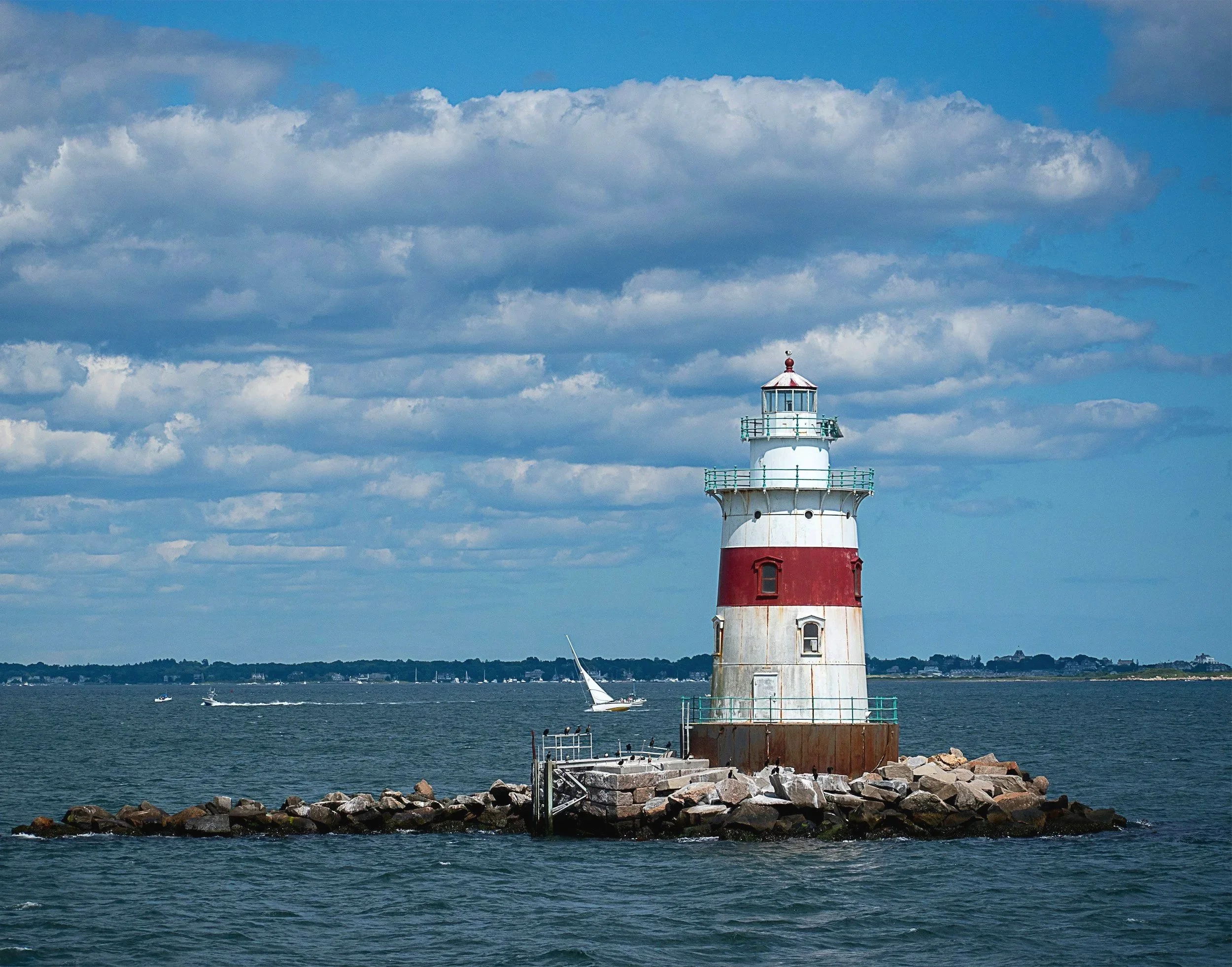 A lighthouse on a rocky breakwater with boats sailing on the water and a cloudy sky in the background.