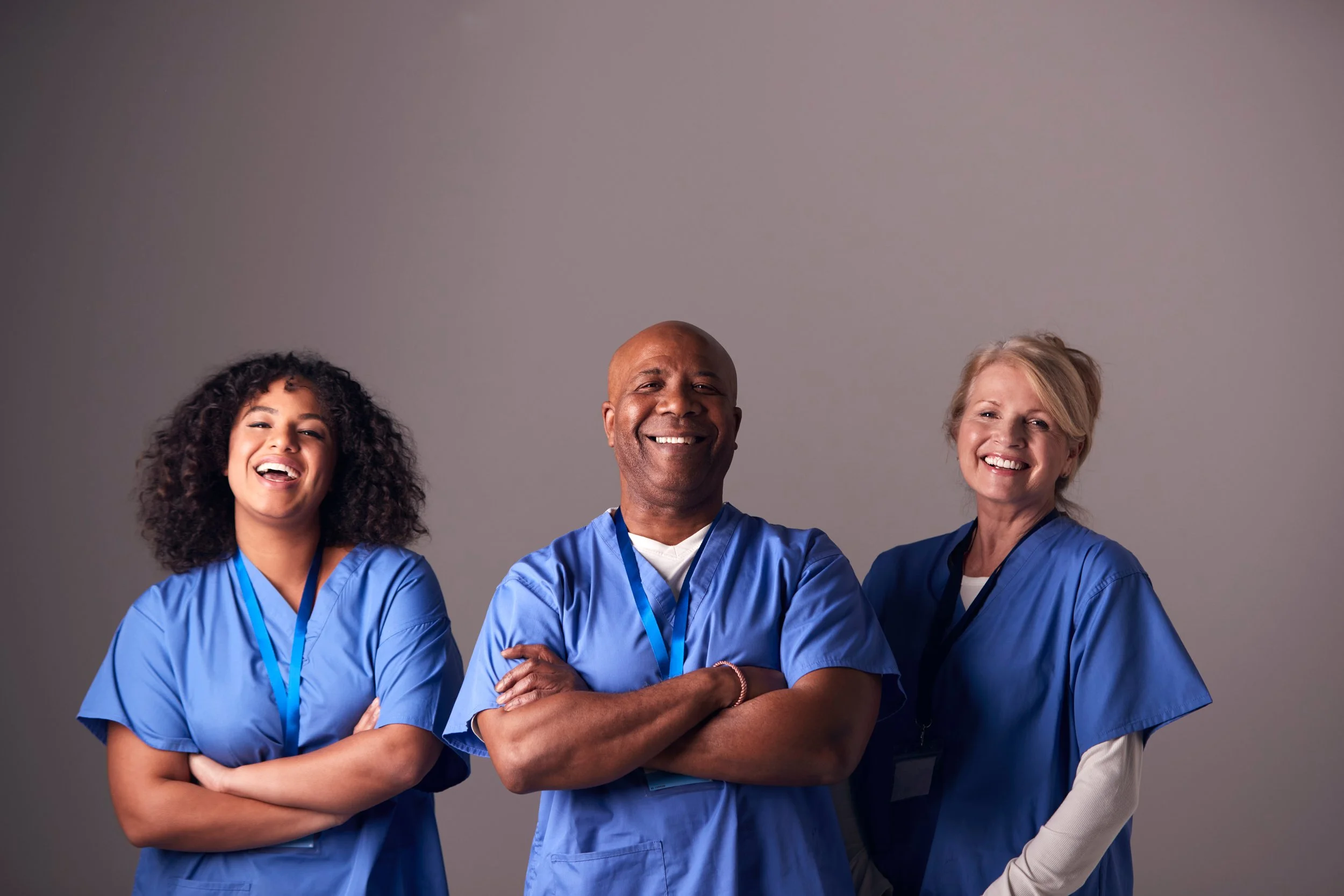 Three healthcare professionals wearing blue scrubs and smiling at the camera against a gray background.