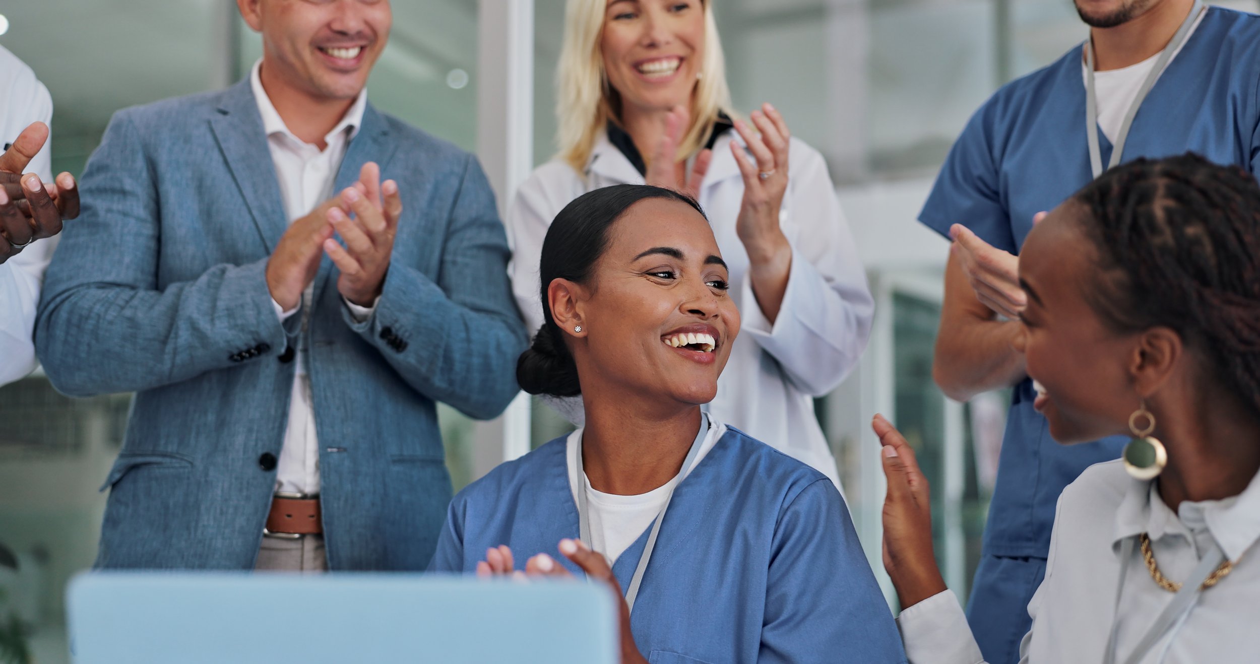 A diverse group of medical professionals celebrating and smiling, with one woman in scrubs sitting at a desk, others standing around, clapping, and engaging with her in a hospital or clinic setting.