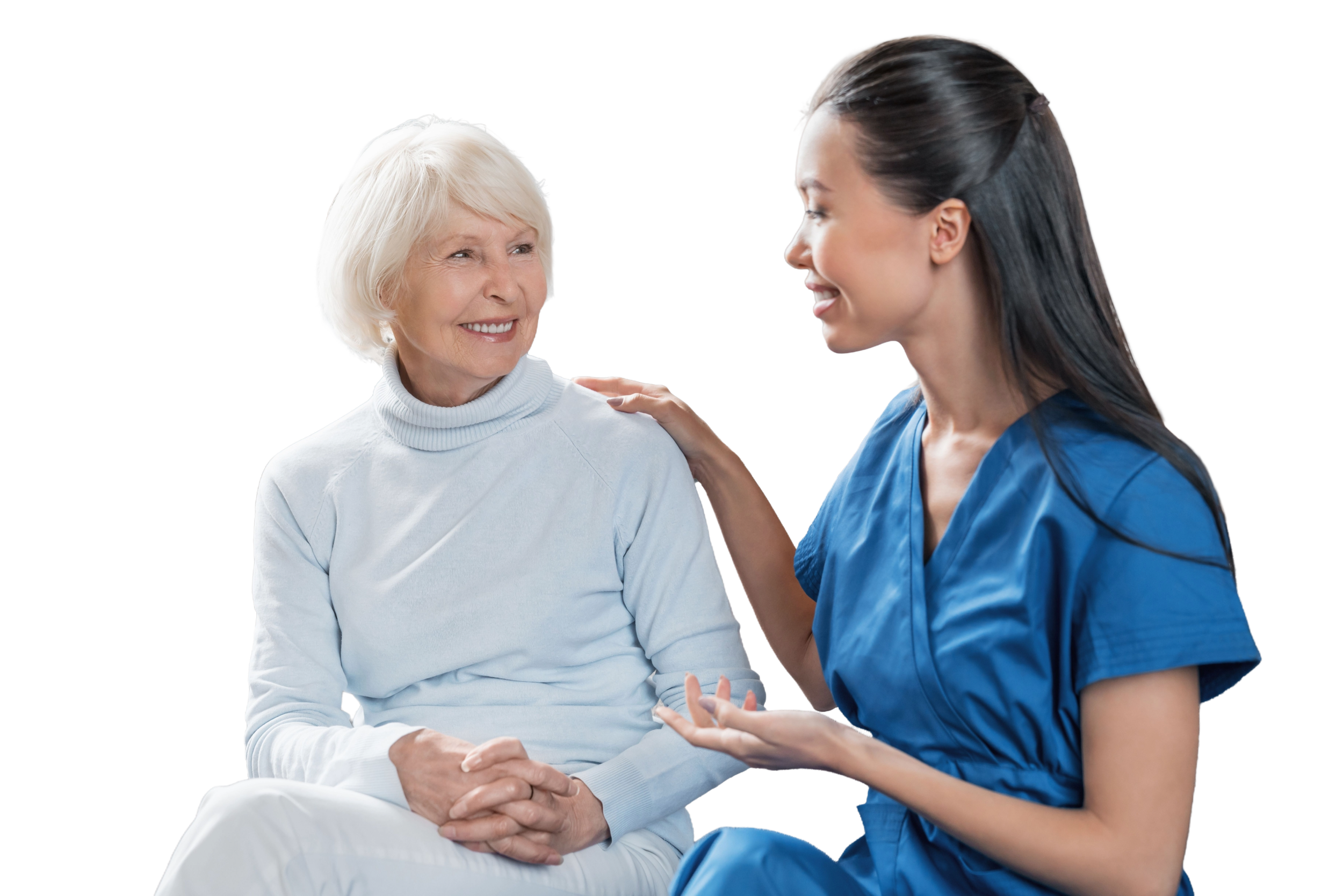 A young female nurse in blue scrubs speaking with an elderly woman in a white turtleneck. They are smiling and having a friendly conversation.