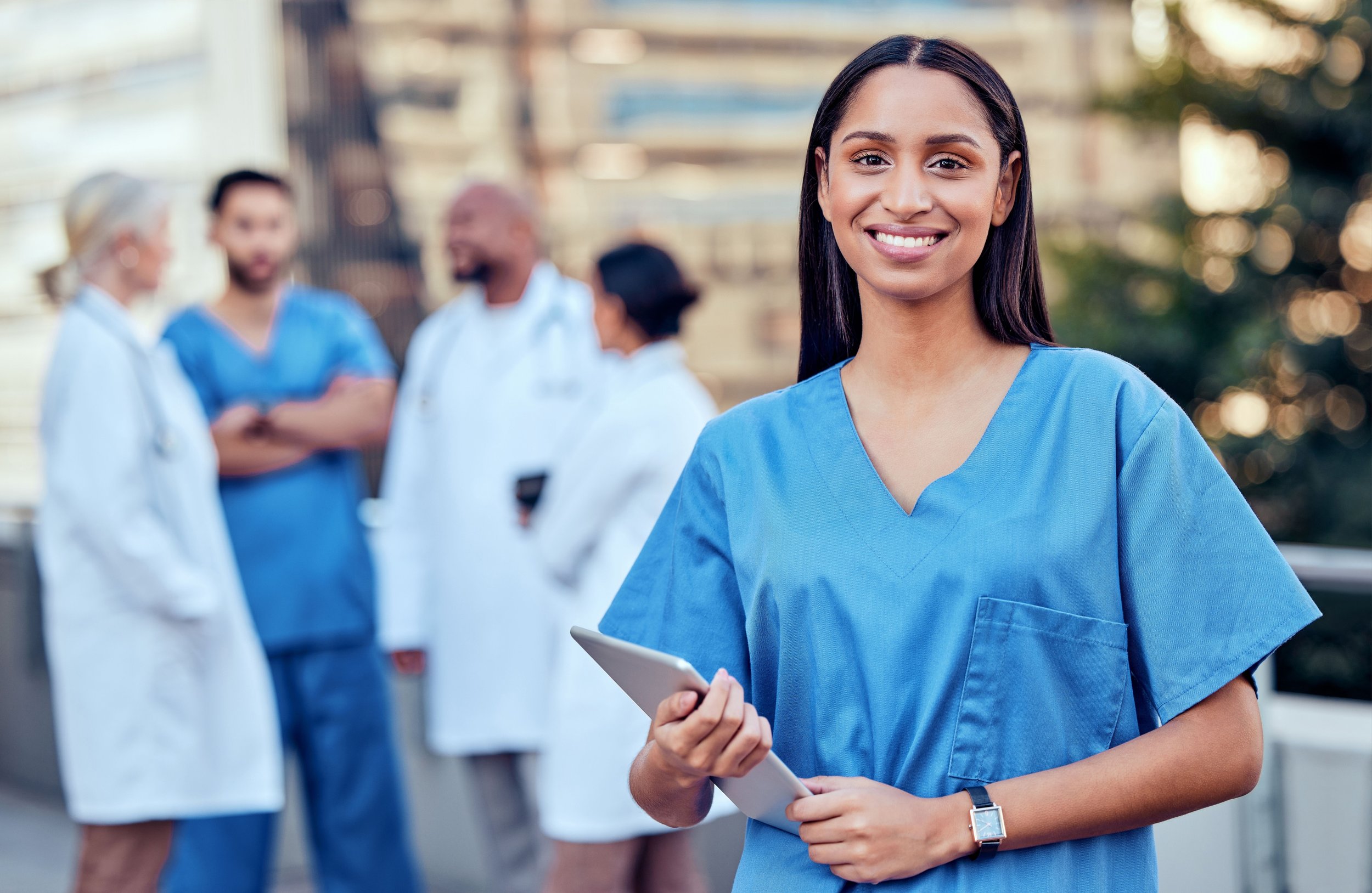 Young nurse wearing blue scrubs smiling at camera, holding a tablet device, with a group of healthcare professionals talking in the background outside in an urban setting.