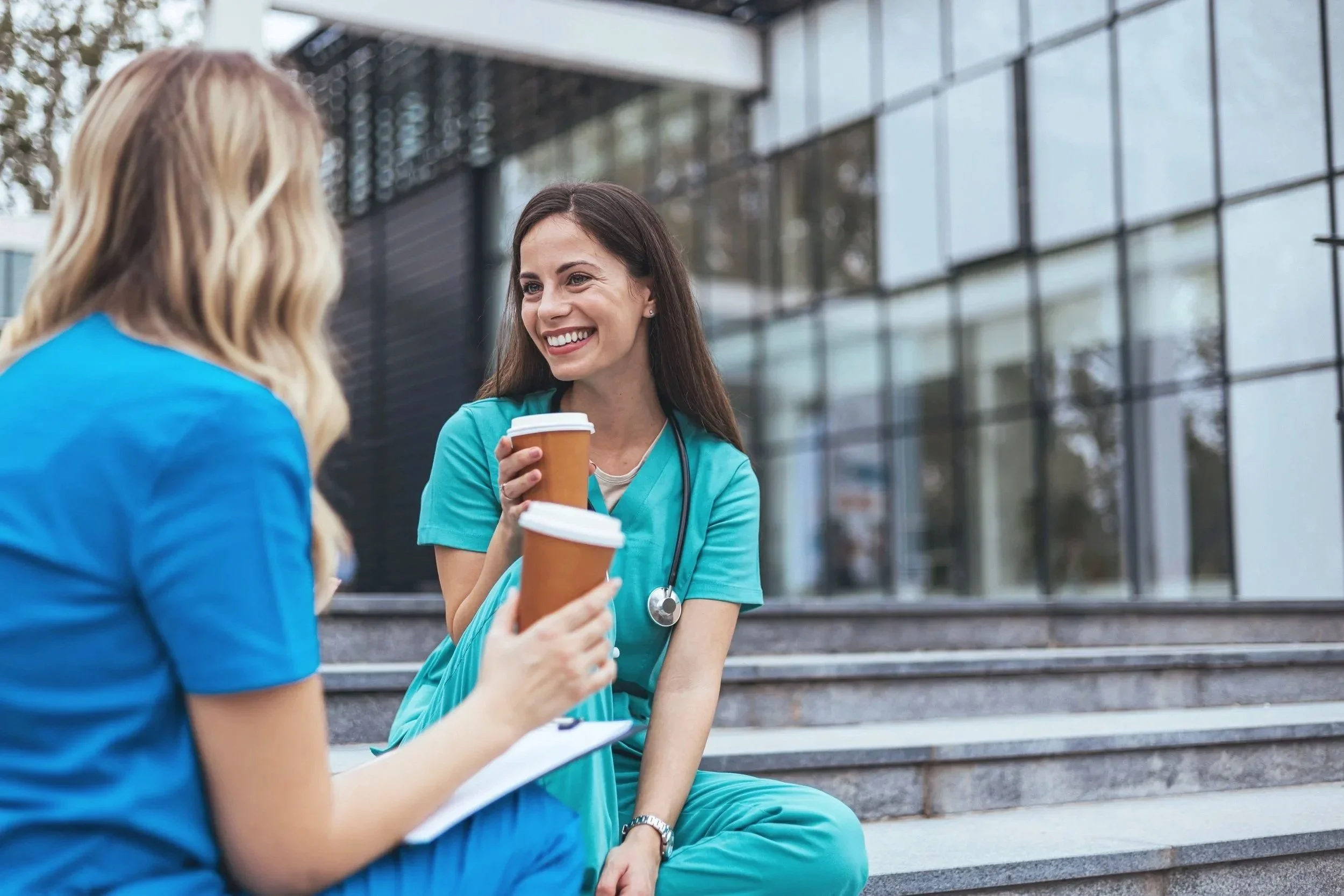 Two female healthcare workers in blue scrubs sitting on outdoor stairs, smiling and chatting while holding coffee cups, outside a modern building with glass windows.