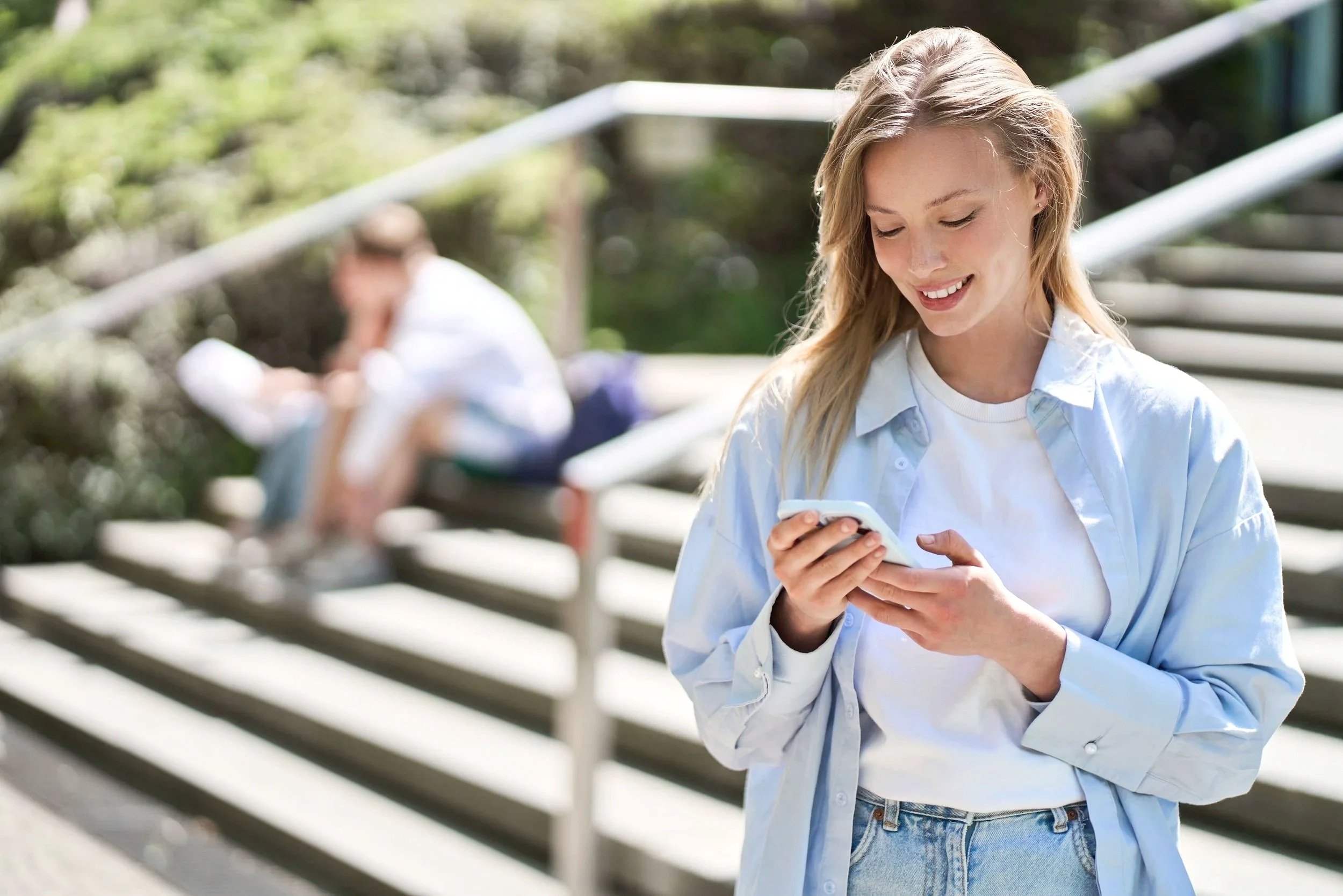 A young woman with long blonde hair smiling while looking at her phone outdoors, with a young man sitting on benches in the background.