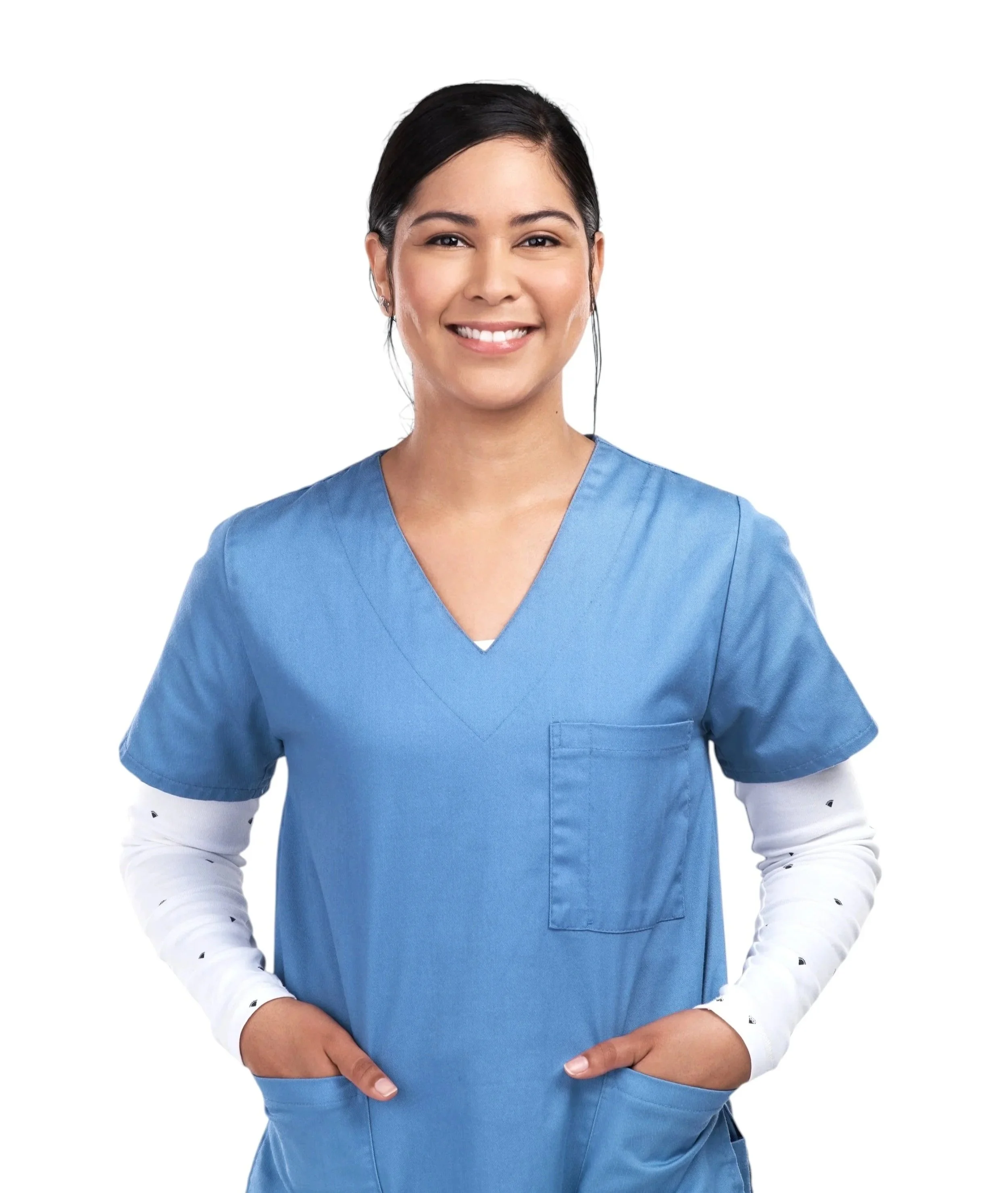 smiling female nurse wearing blue scrubs and white long sleeve shirt with small black dots, hands in pockets, against white background