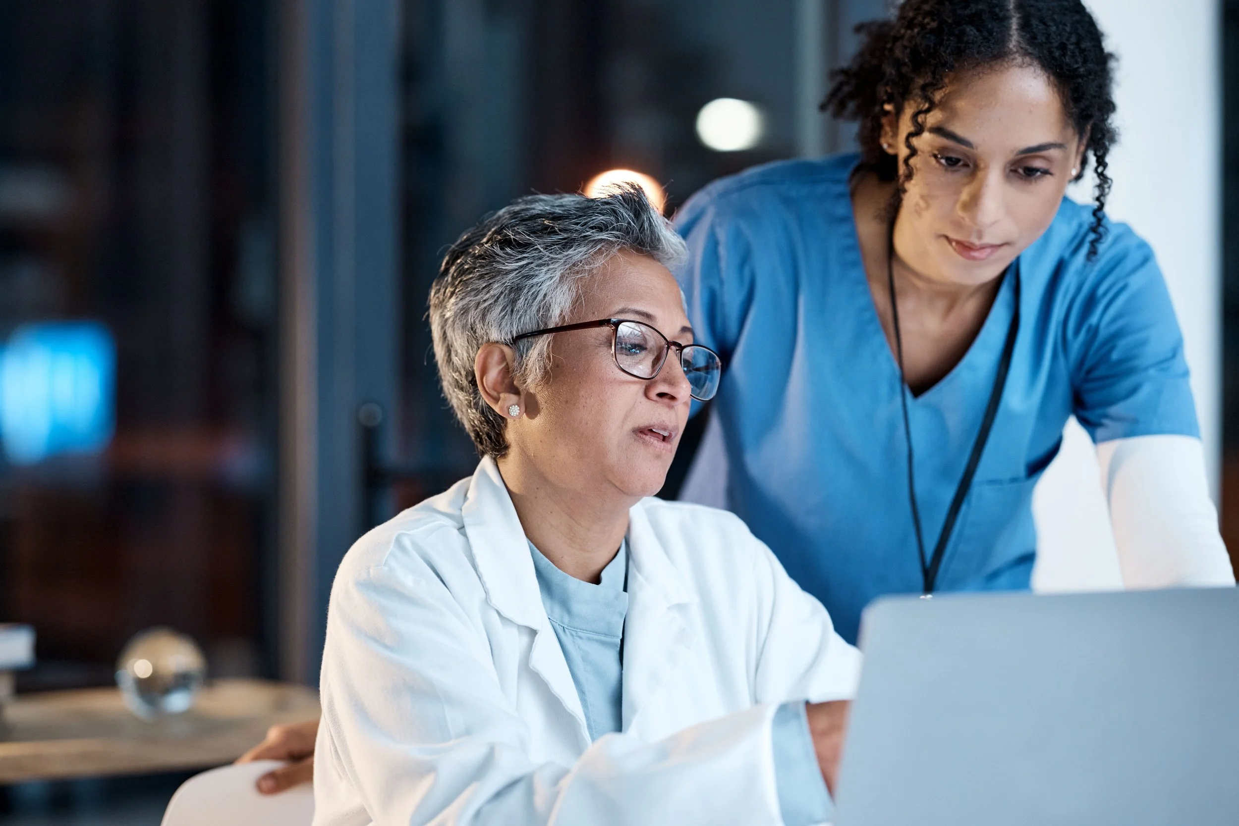 Two female healthcare professionals, one older with glasses and gray hair in a white coat, and the other younger with curly dark hair in blue scrubs, working together on a laptop in a modern medical facility.