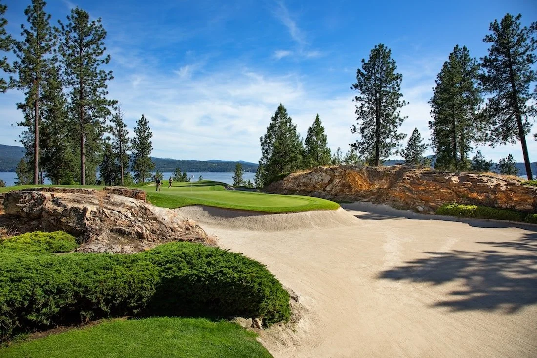 Sand, rock outcroppings, and trees surround a green being played by two golfers at the Coeur d’Alene Resort Golf Course, with the lake in the background.