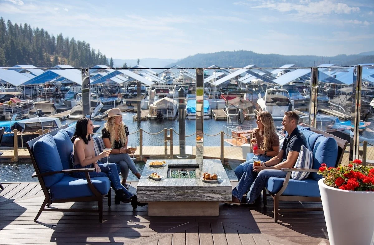 Four guests on the Whispers Lounge patio enjoying drinks and light bites around a fire pit, with docked boats and Lake Coeur d’Alene behind them.