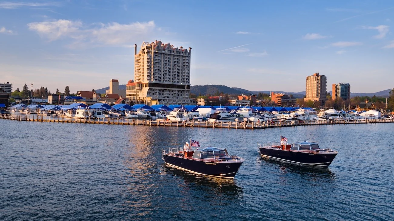 Two classic wooden boats on the water with the Coeur d’Alene Resort Marina and Lake Tower in the background.