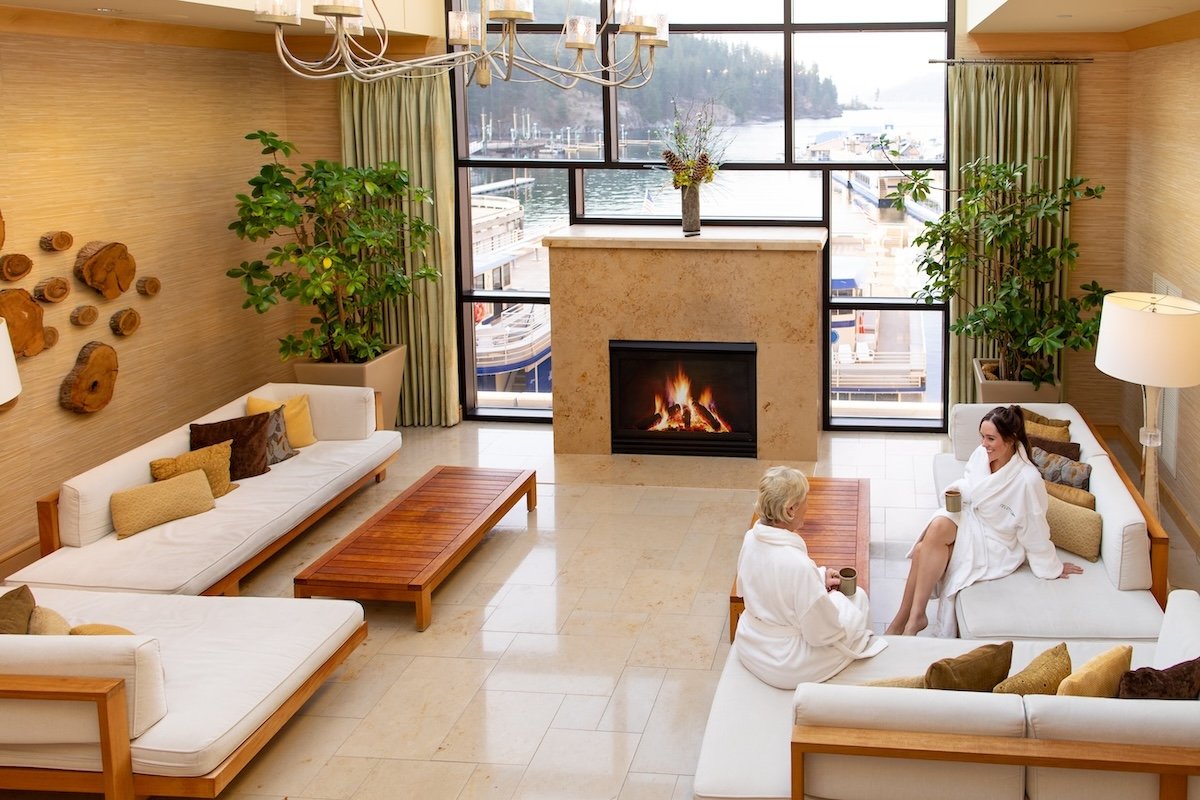 Two women relaxing in robes in the Coeur d’Alene Resort spa lounge with a lit fireplace and views of the dock and Lake Coeur d’Alene.