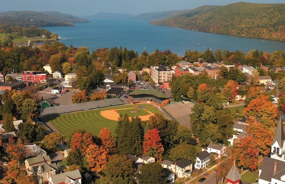 An empty baseball stadium centers a Cooperstown aerial view, fall foliage and the lake beyond.