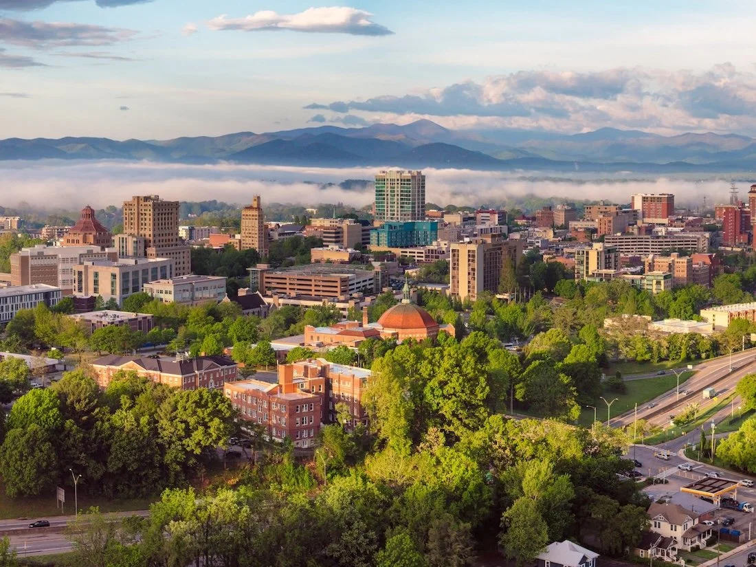 Aerial view of Downtown Asheville with clouds hovering by the city skyline and the Blue Ridge Mountains in the distance.