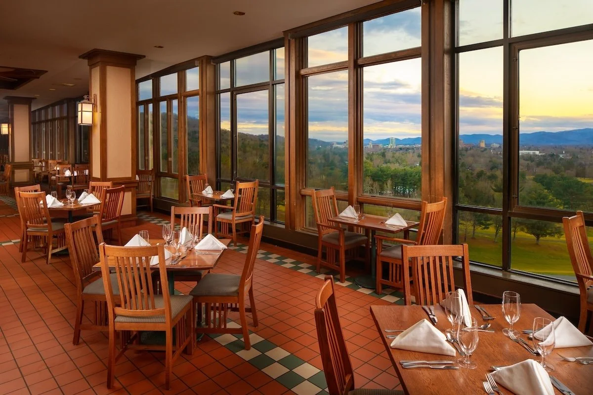 Set tables near floor-to-ceiling windows overlooking trees, Asheville, and mountains in the Grove Park Inn’s Blue Ridge dining room.