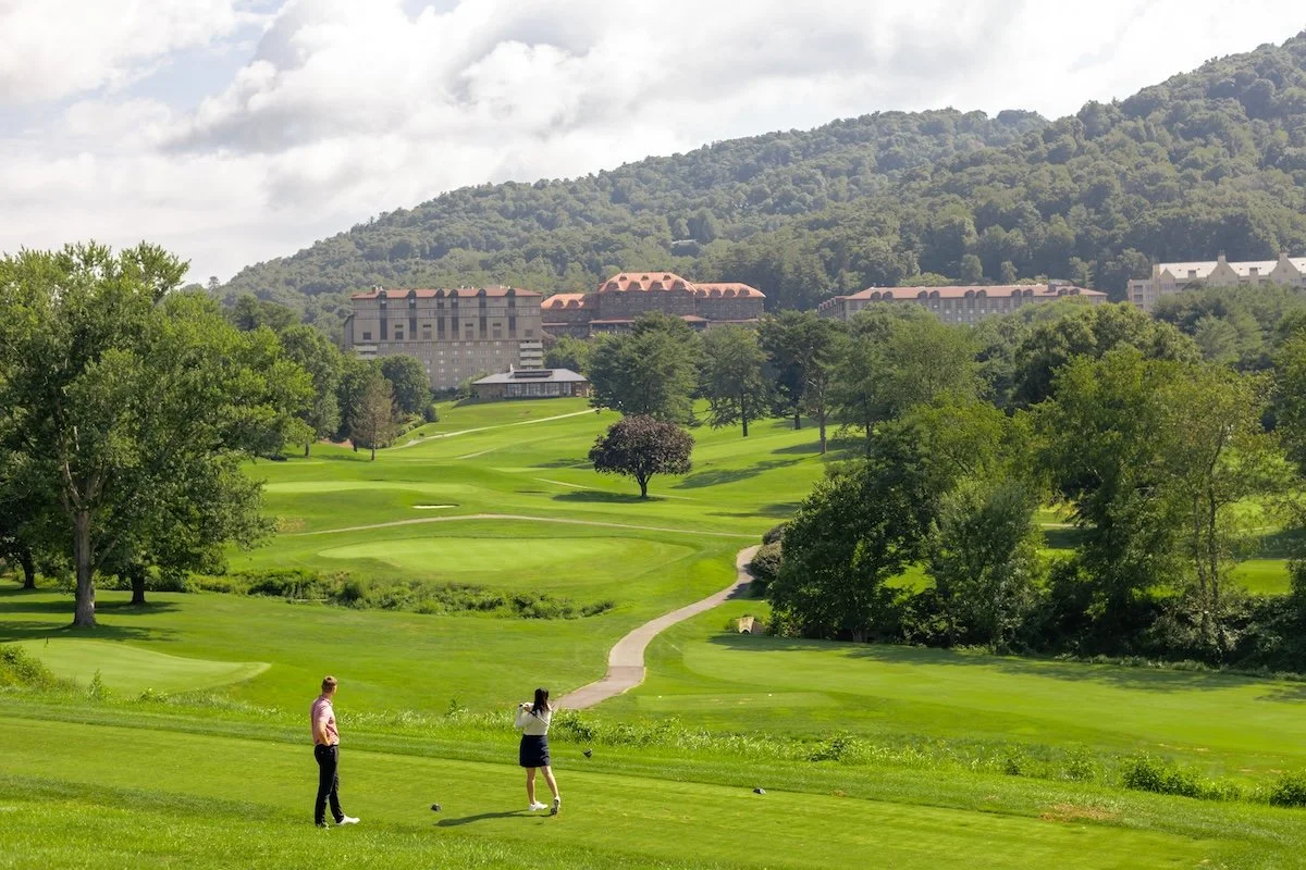Male golfer watching a female golfer tee off on a par-three hole at the Omni Grove Park Inn Golf Course with the Inn in the background.